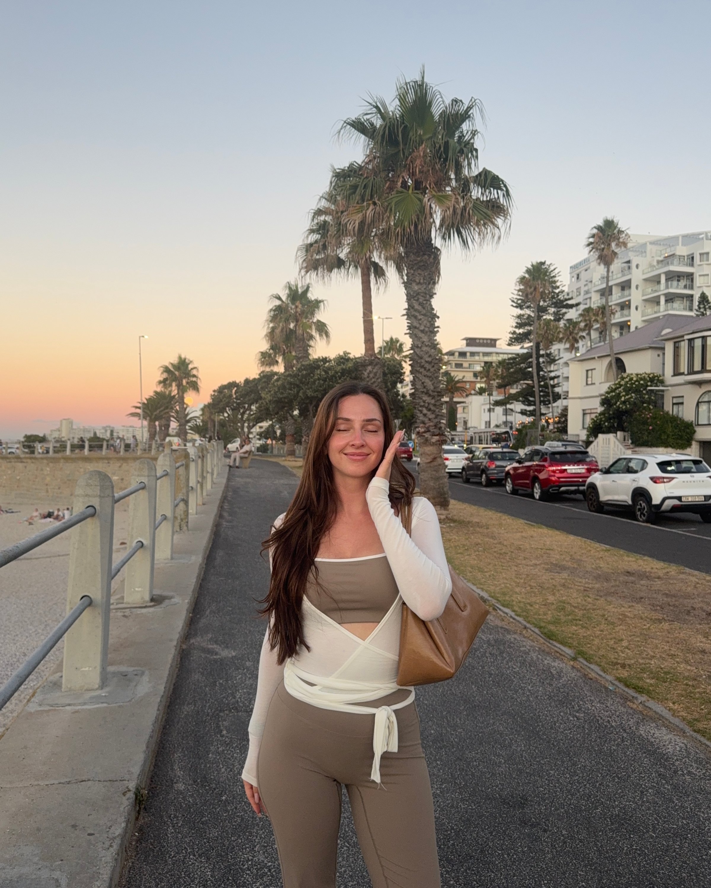 A woman stands on a seaside path during sunset, wearing a stylish outfit and holding a bag, with palm trees and buildings in the background.