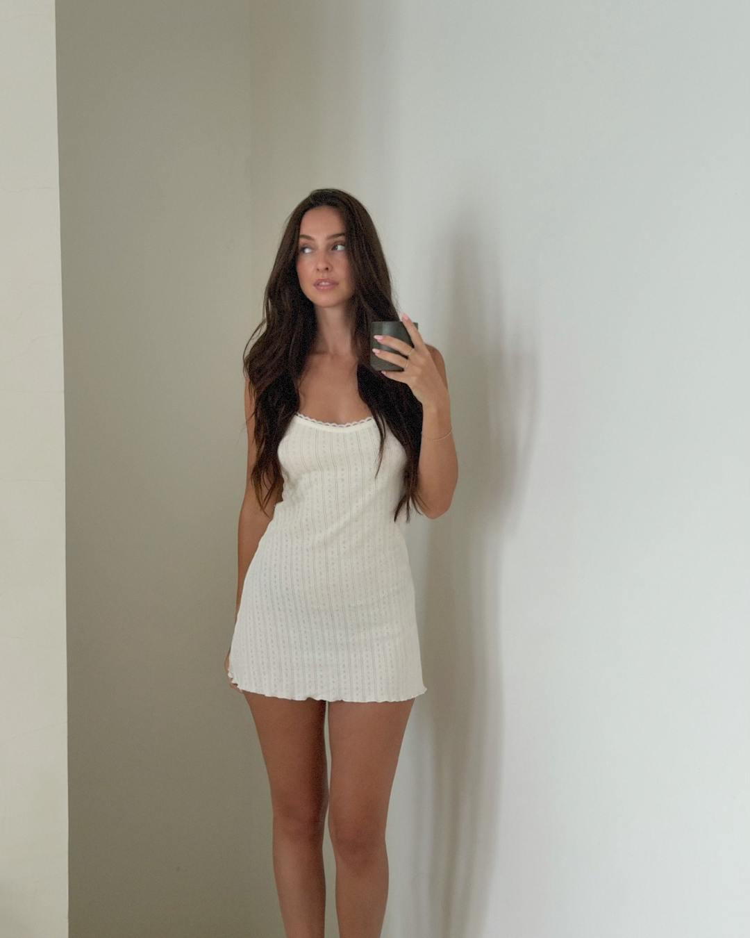 A woman stands against a plain wall, wearing a white dress and holding a phone. She has long, dark hair and a natural expression.