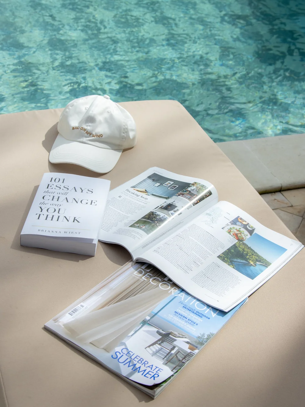 A relaxing scene featuring a book titled '101 Essays That Will Change the Way You Think', a magazine open to a page about decoration, and a white cap, all placed on a sunbed beside a swimming pool.