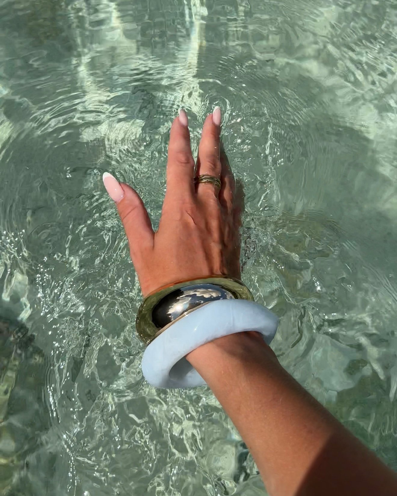 A hand with manicured nails adorned with multiple bracelets, reaching into clear water.