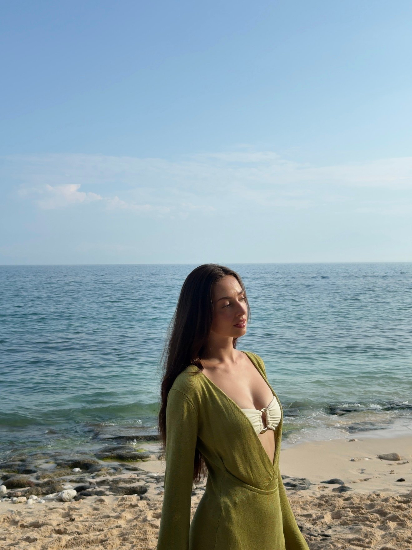 A woman stands on a beach, gazing thoughtfully towards the ocean, wearing a green cover-up over a bikini.