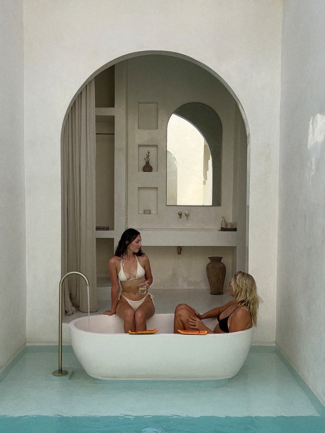 Two women relax in a modern bathtub surrounded by minimalist architecture and water features.