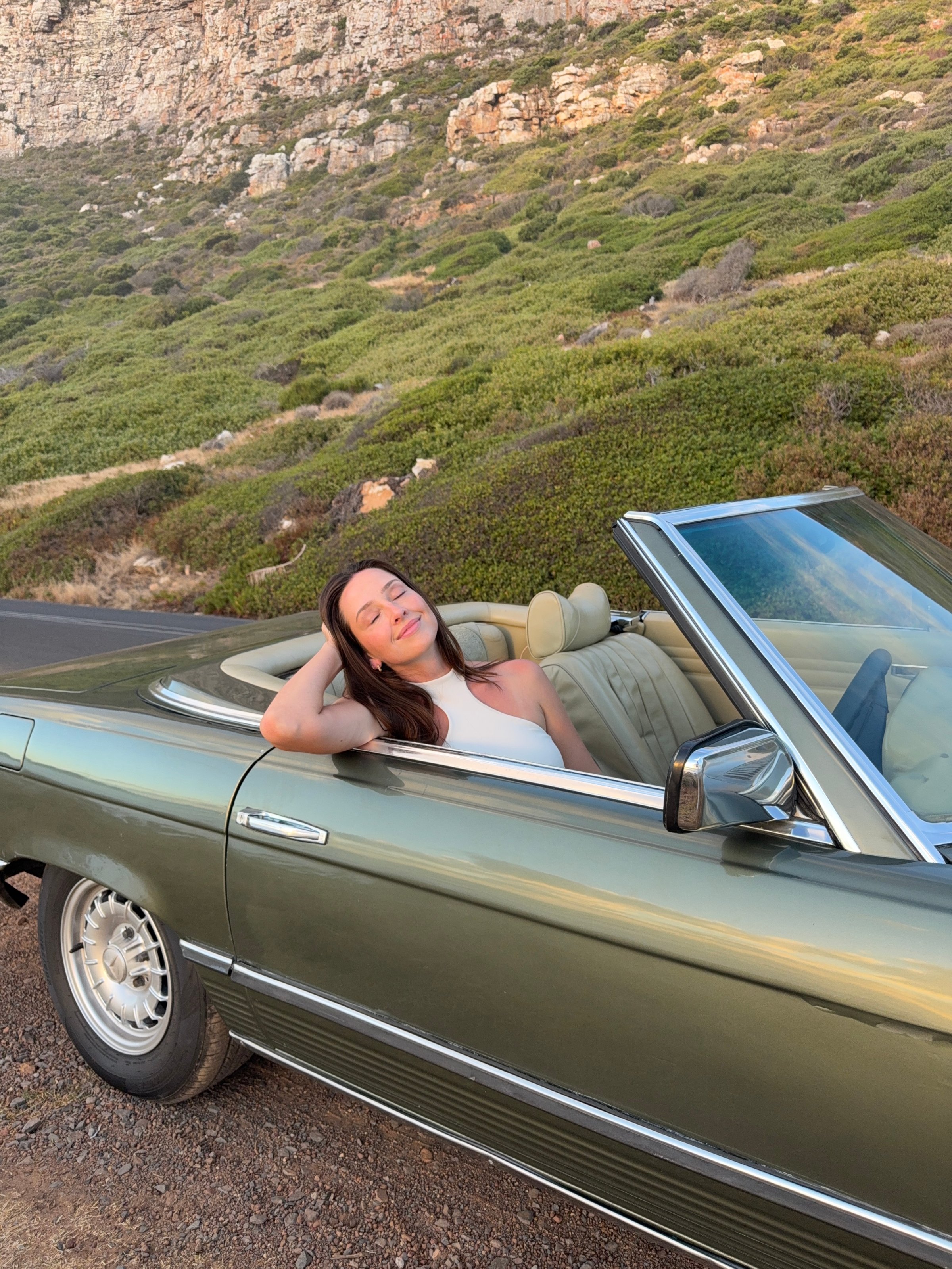 A woman relaxes in a convertible car parked on a scenic road surrounded by greenery and rocky cliffs.