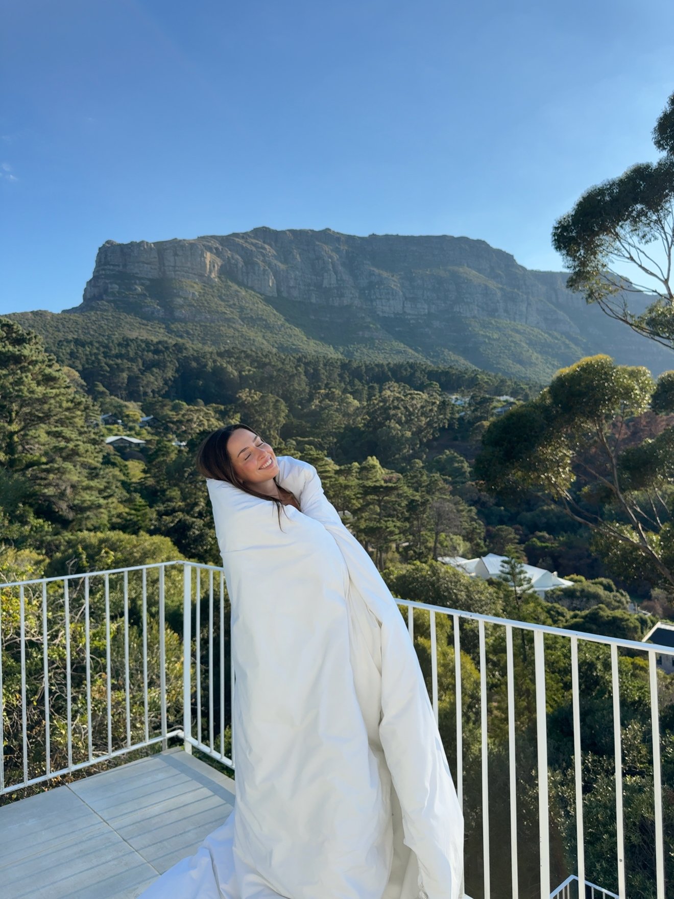 A woman wrapped in a white blanket stands on a balcony with a scenic mountain view in the background, smiling joyfully.