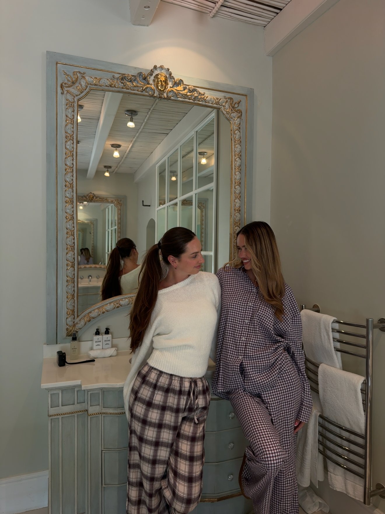 Two women in cozy outfits pose together in a stylish bathroom with a large mirror and elegant decor.