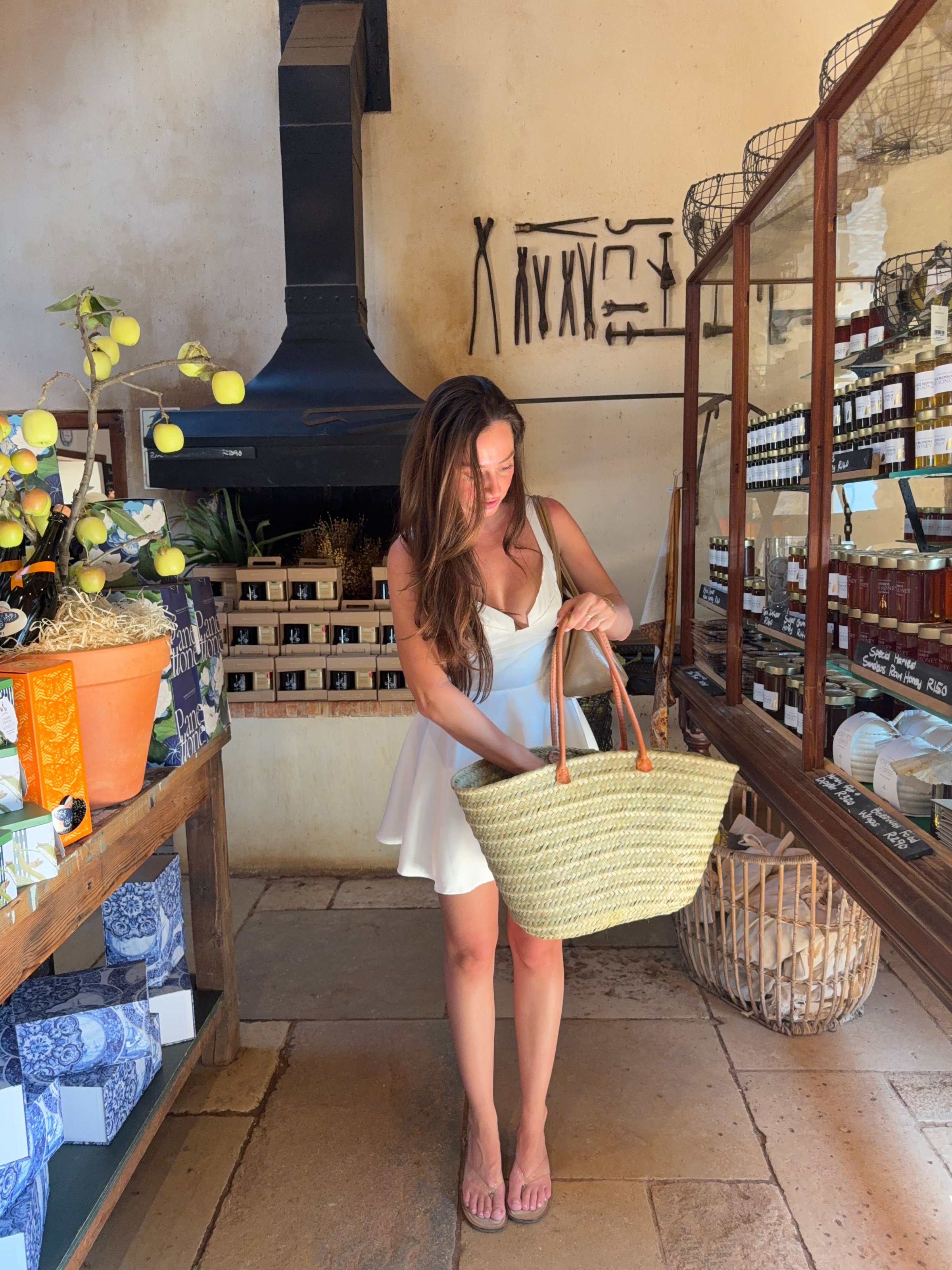 A woman in a white dress stands inside a shop, holding a large woven bag while surrounded by various products and decorations.