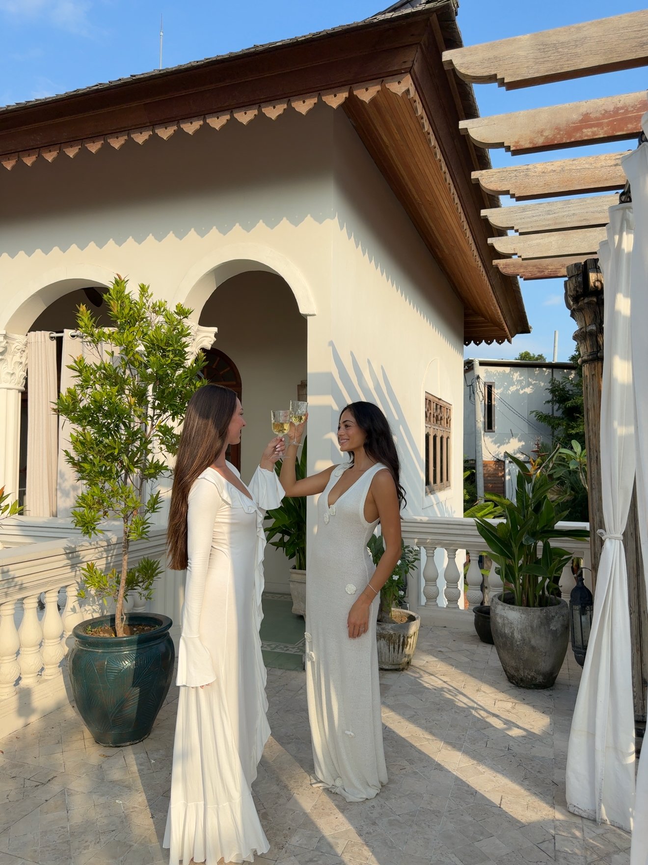 Two women in elegant dresses toast with glasses in a beautifully designed outdoor setting.