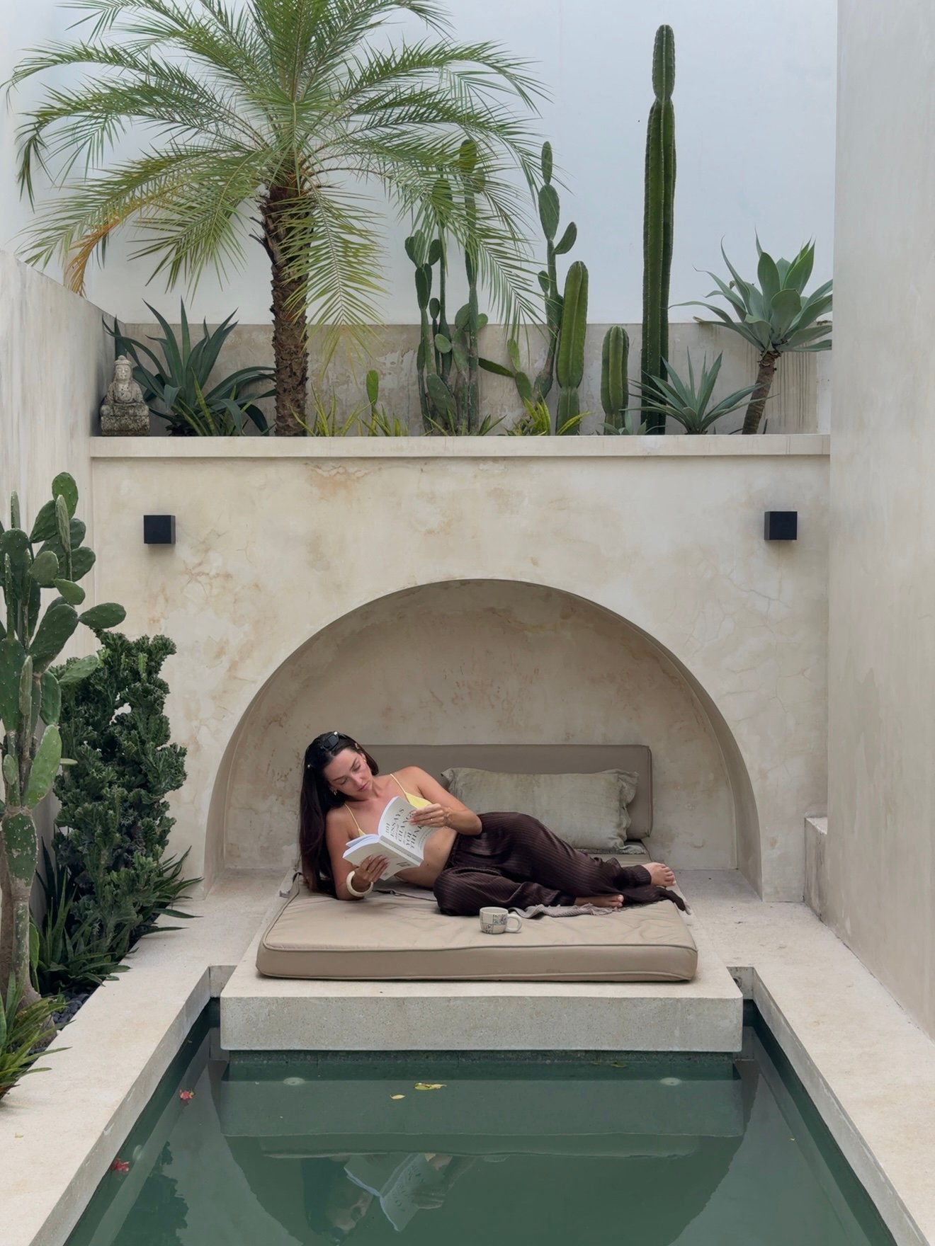 A woman relaxes on a lounge area beside a serene pool, surrounded by lush greenery and modern architecture, while reading a book.