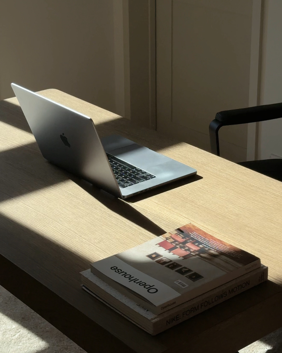 A sleek laptop sits on a wooden desk, illuminated by natural light, with a book titled 'Opensource' placed beside it.