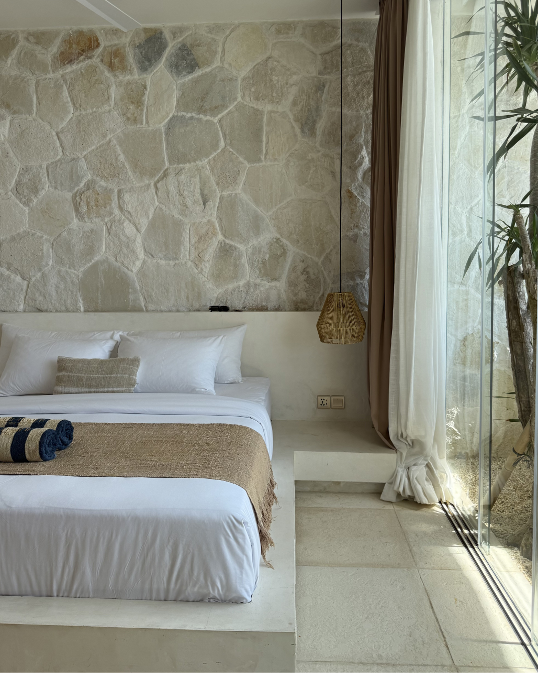 A serene bedroom featuring a stone wall, a cozy bed with white linens, and a woven pendant light. Natural light streams in through large glass windows, highlighting the minimalist decor.