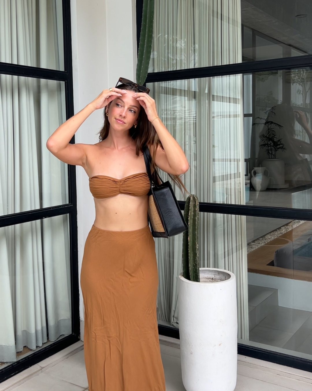 A woman stands in a modern indoor setting, wearing a brown two-piece outfit and holding sunglasses. A tall cactus is visible in the background.