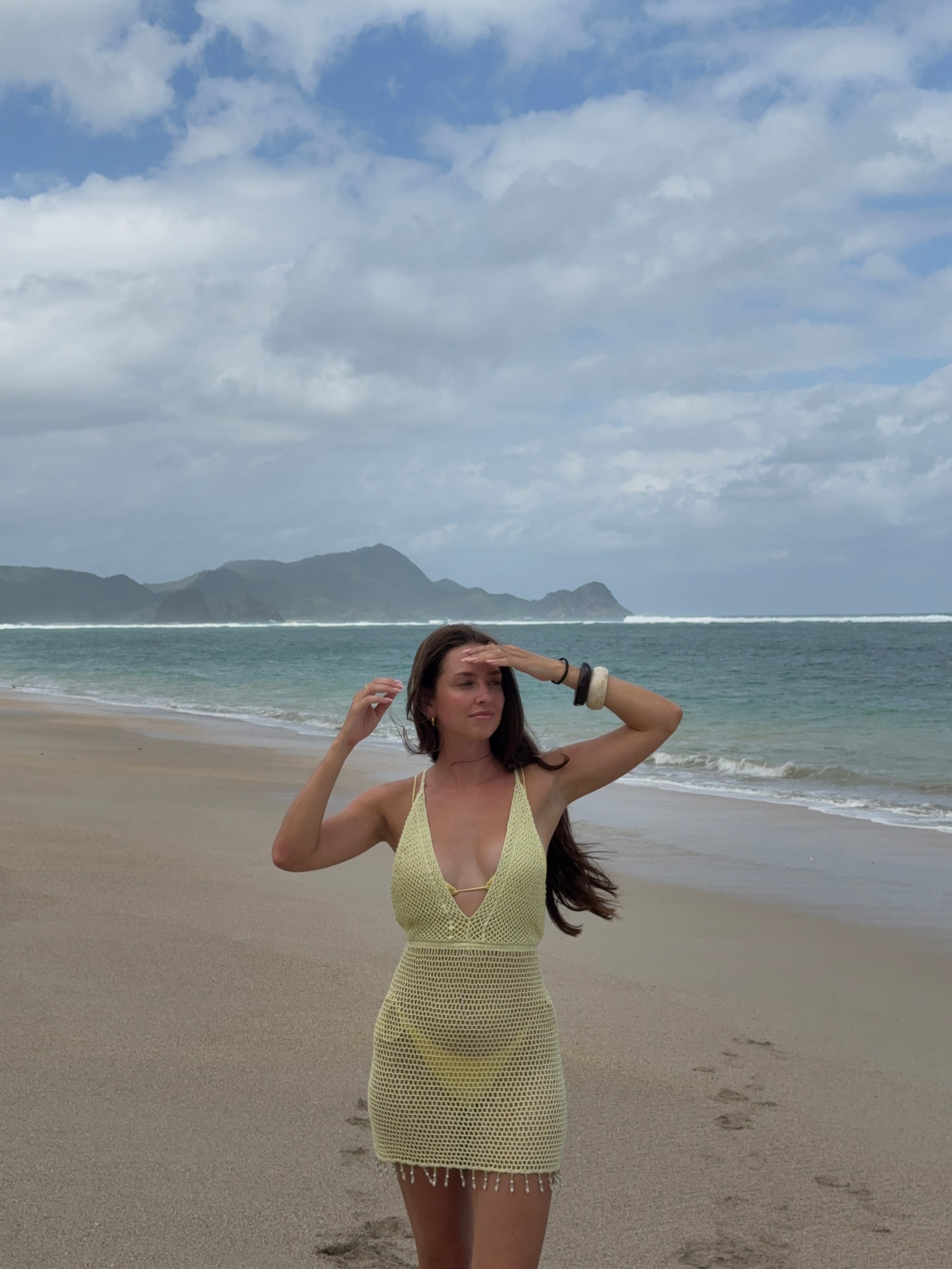 A woman in a yellow crochet dress stands on a sandy beach, shielding her eyes from the sun while looking out at the ocean and distant mountains.