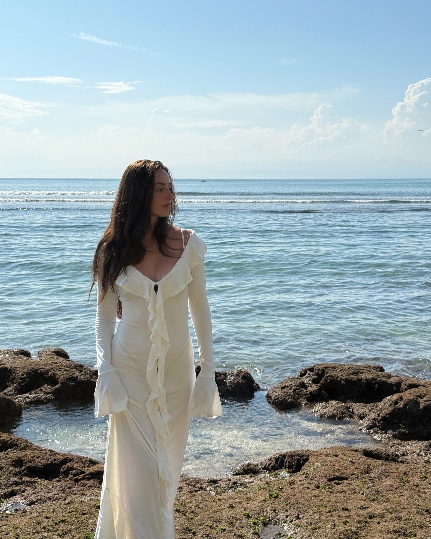 A woman in a white dress stands on rocky shorelines by the sea, with a serene ocean and cloudy sky in the background.