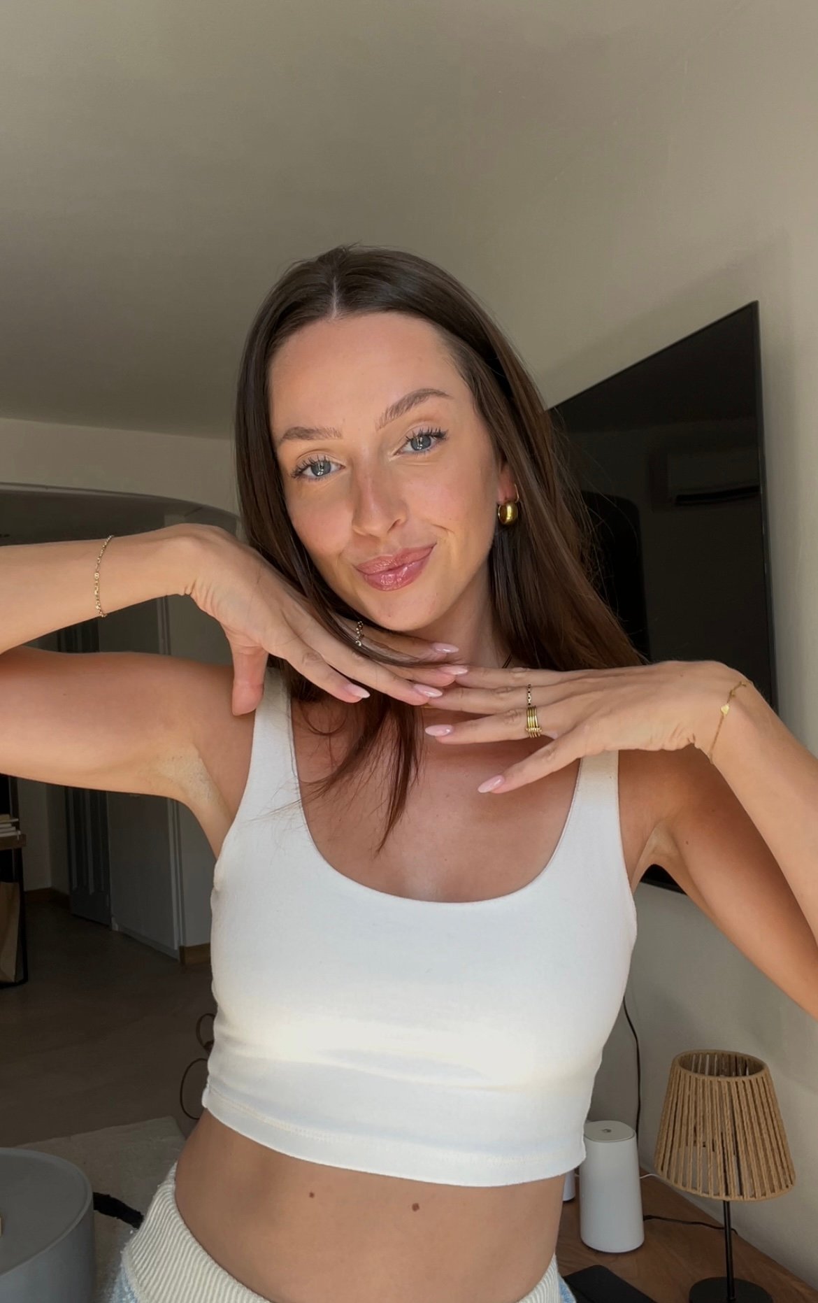 A woman with long brown hair poses playfully in a bright indoor setting, wearing a white tank top and showcasing her jewelry.