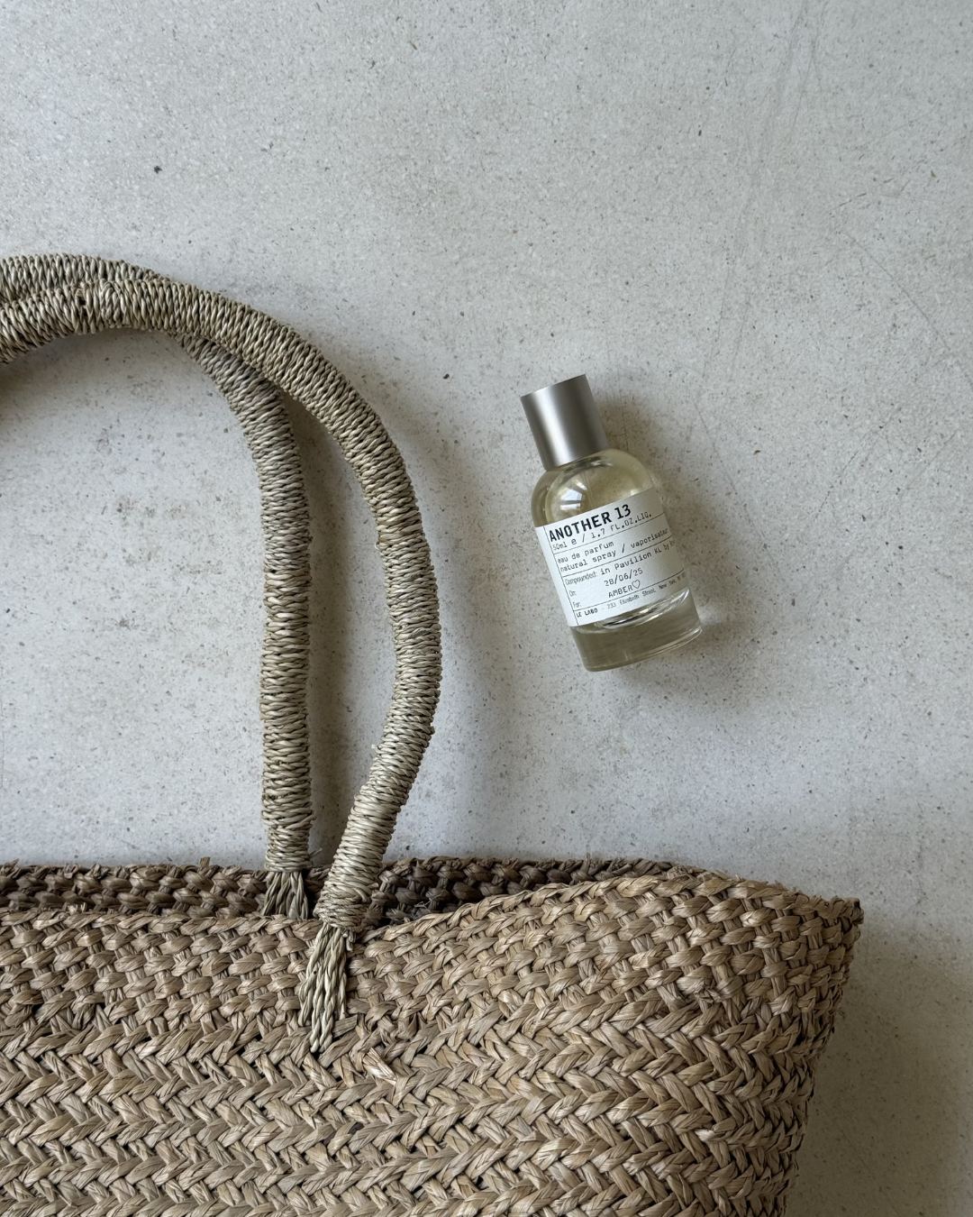 A woven straw bag next to a bottle of fragrance, showcasing a minimalist aesthetic.