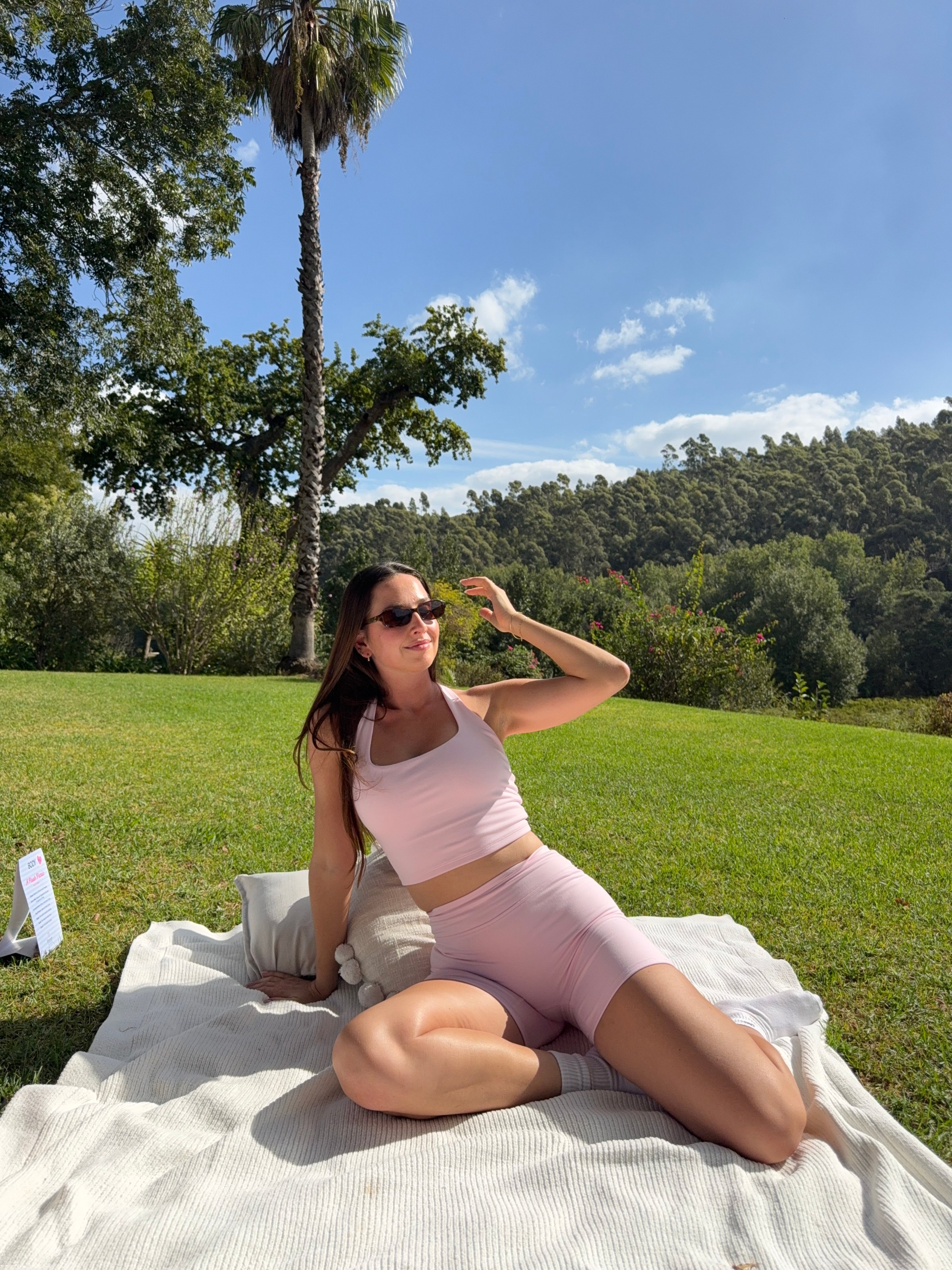 A woman in a pink athletic outfit poses on a blanket in a grassy area, with trees and a blue sky in the background.