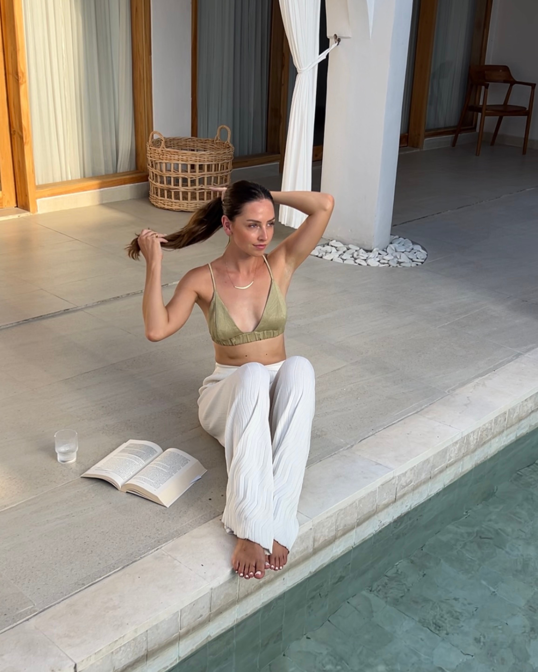 A person sitting by a pool, wearing a green top and white pants, with a book in hand and hair being styled.