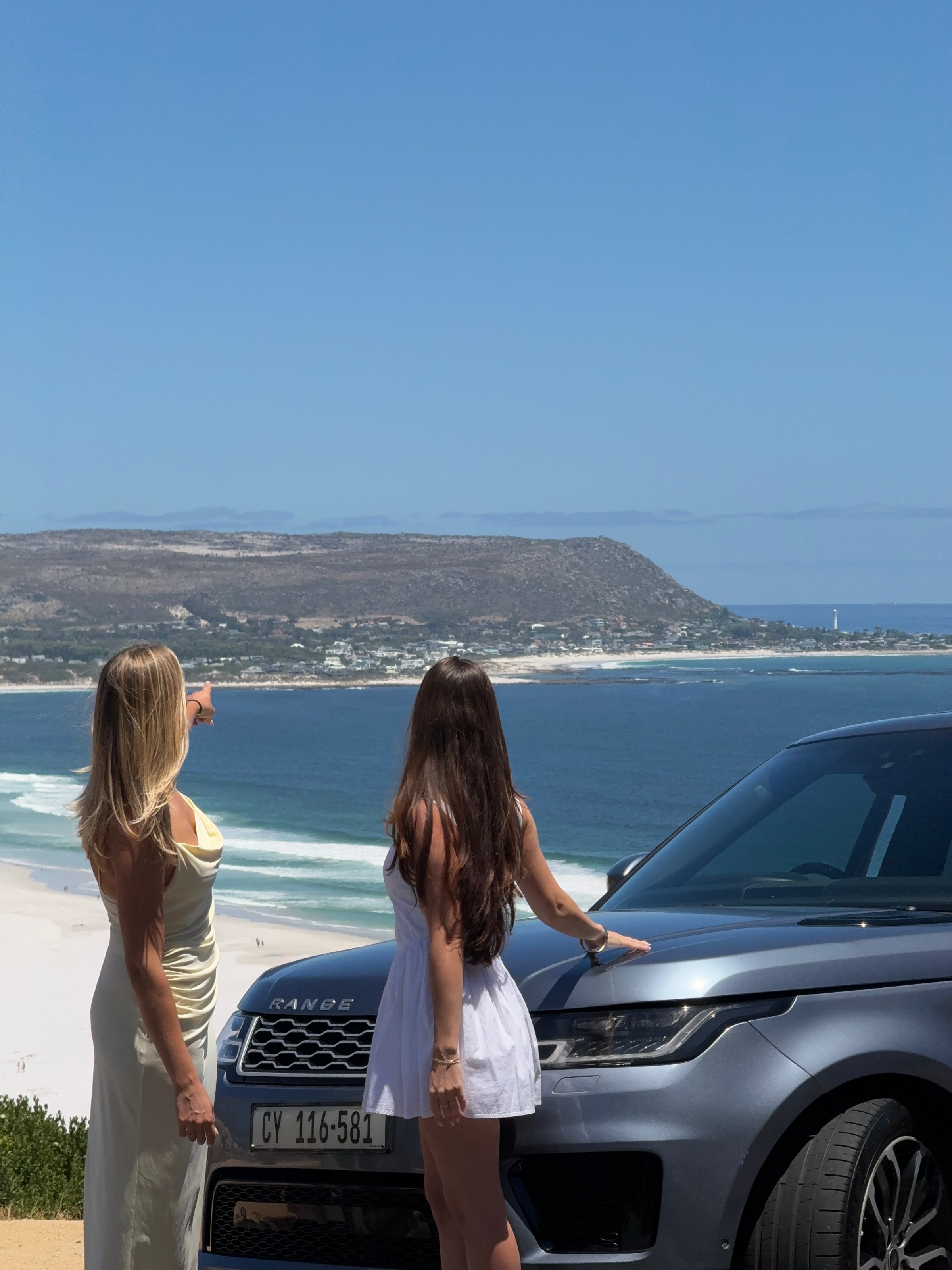 Two women standing beside a Range Rover, pointing towards the ocean and coastline in the background on a sunny day.