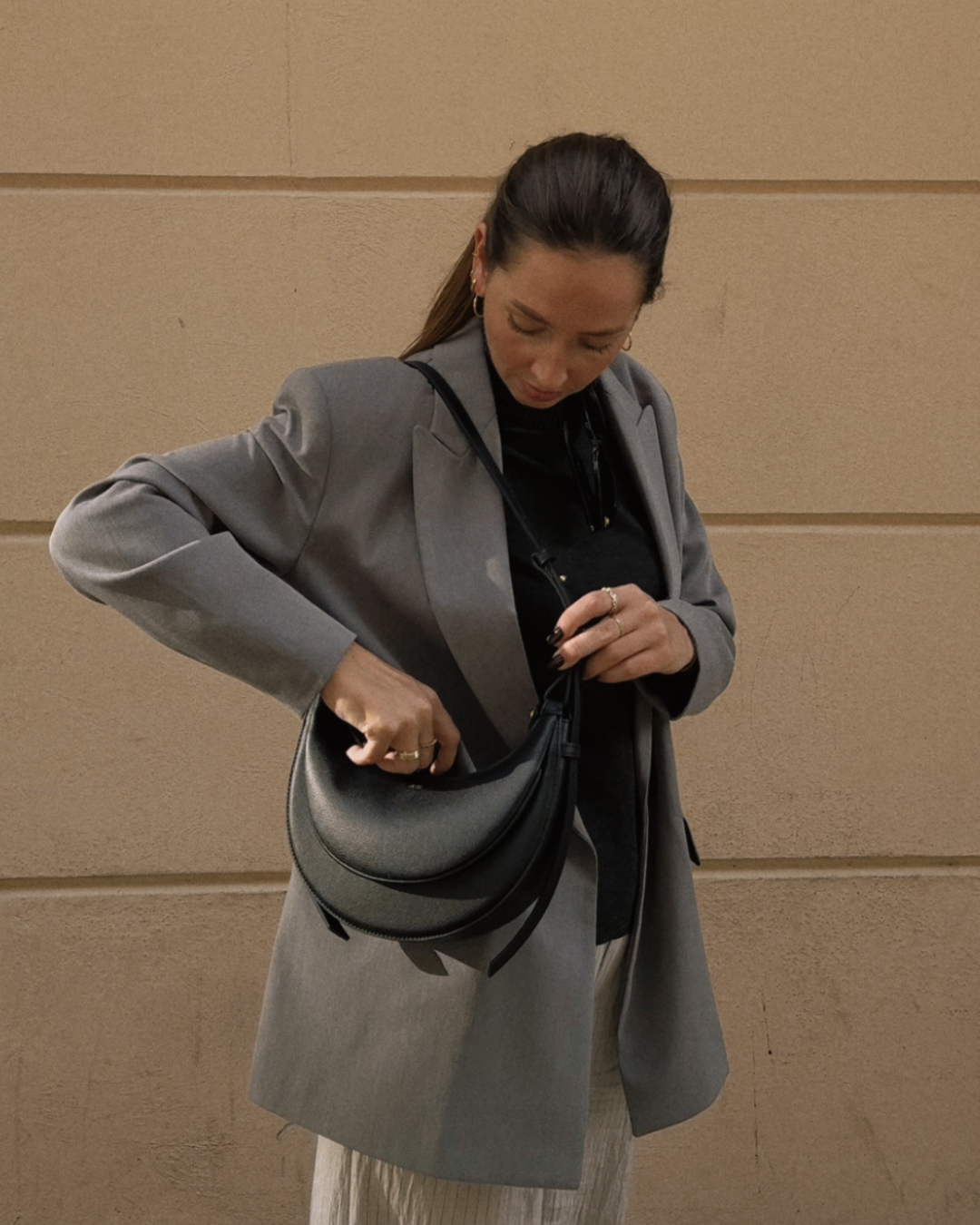 A woman in a gray blazer adjusts a black handbag while standing against a beige wall.