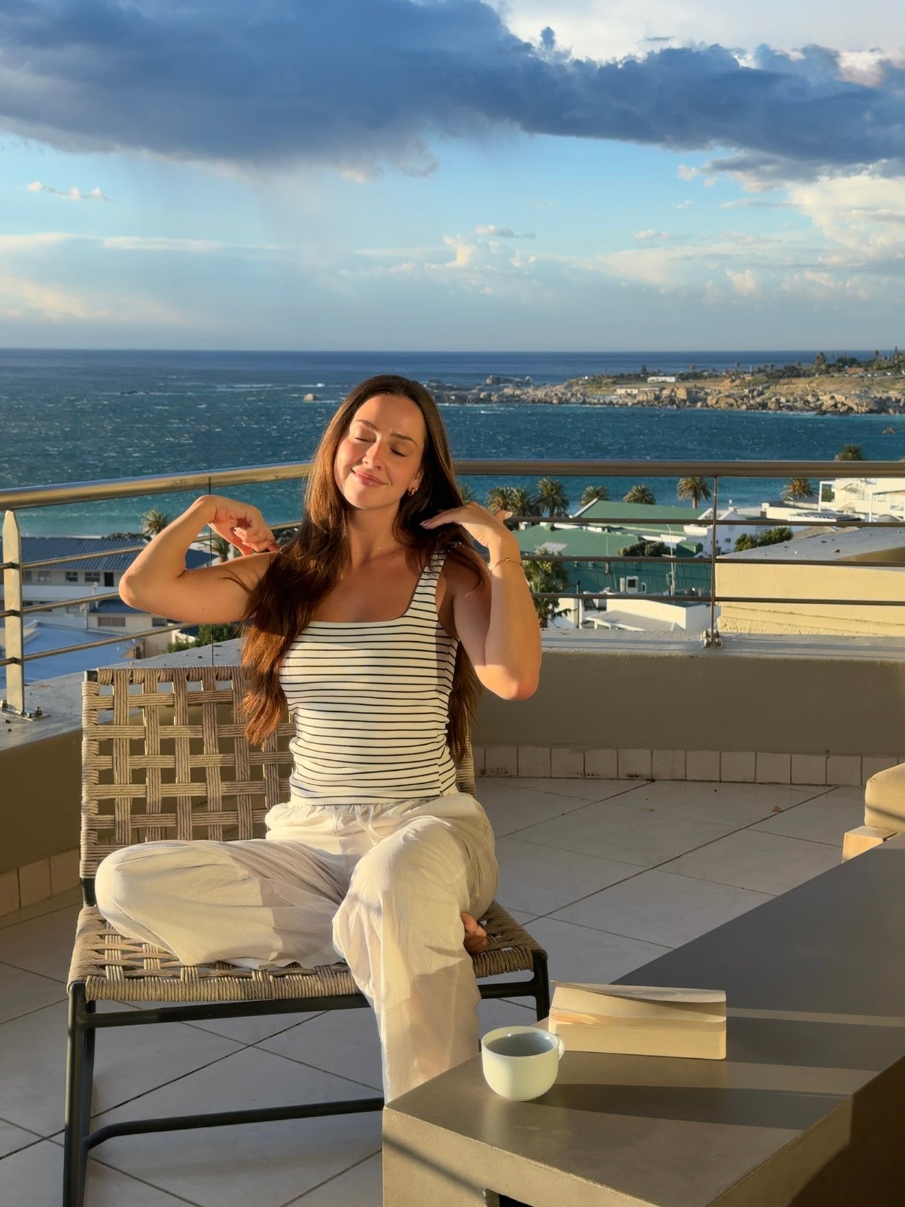 A woman with long hair sits on a chair on a balcony, enjoying the view of the ocean and sky. She wears a striped tank top and light pants, with a relaxed expression.