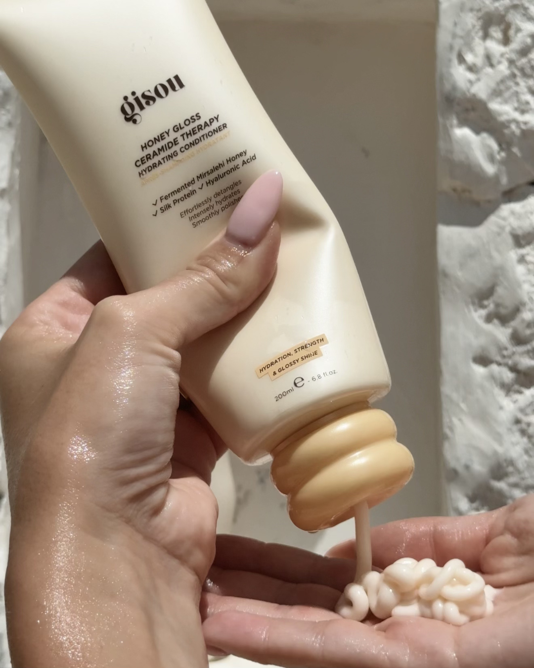 A hand holding a tube of Gisou Honey Gloss Ceramic Therapy Hydrating Conditioner, with the product being dispensed into the palm.