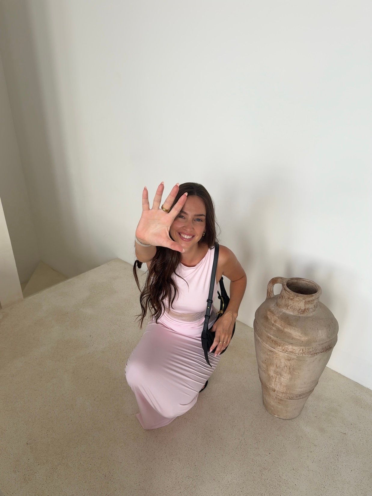 A woman in a pink dress is sitting on the floor, smiling and waving her hand. Next to her is a large clay pot.