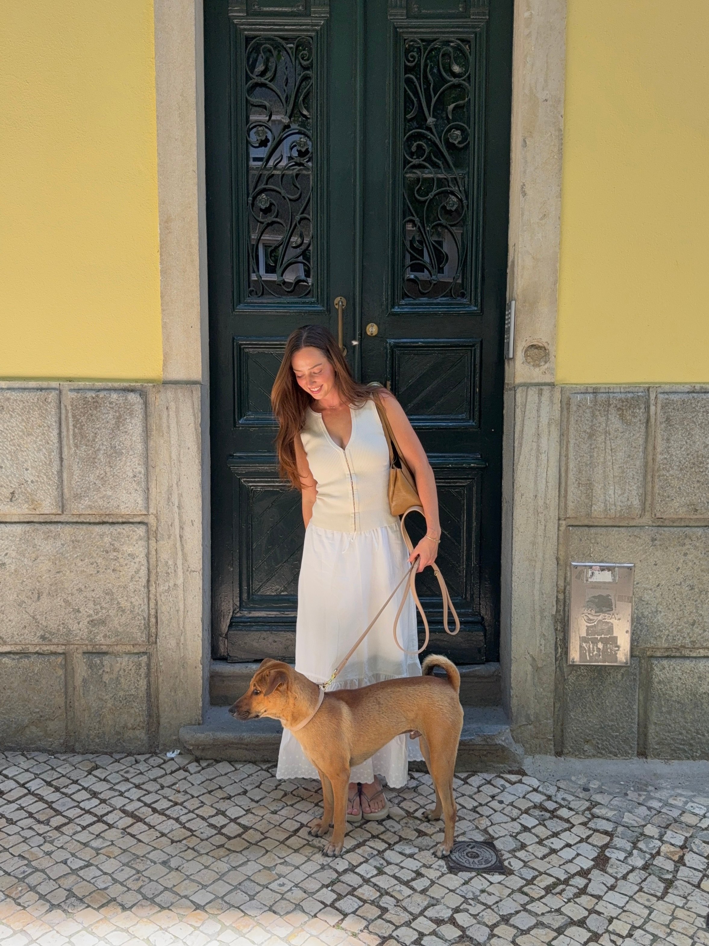 A woman in a white dress stands beside a brown dog in front of a green door with decorative features.