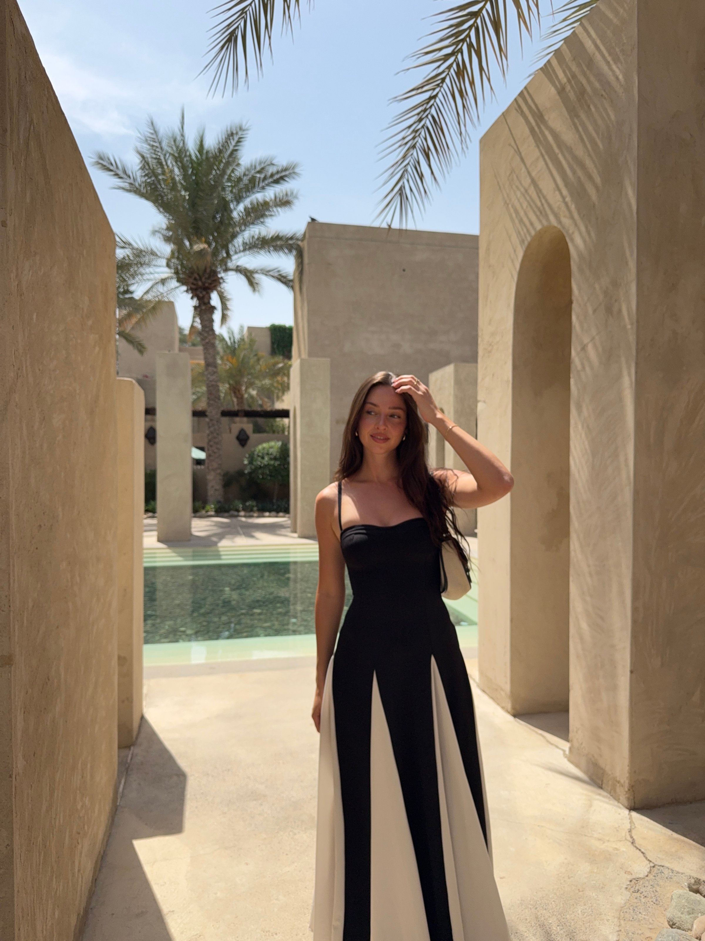 A woman stands in a stylish black and white dress in an outdoor setting with palm trees and a pool in the background.
