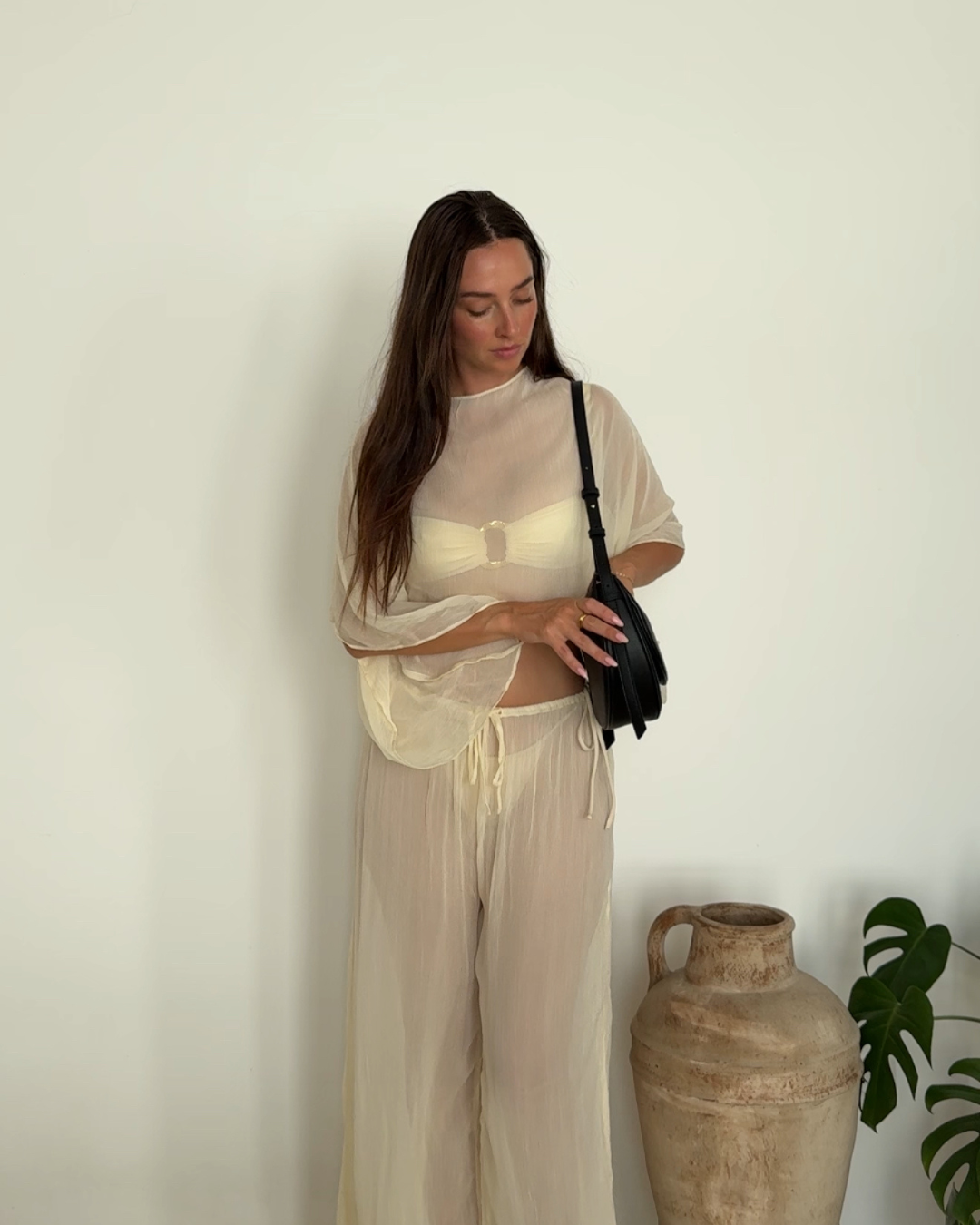 A woman in a sheer, light-colored outfit stands thoughtfully, holding a small black bag. A large clay pot and a green plant are visible in the background.