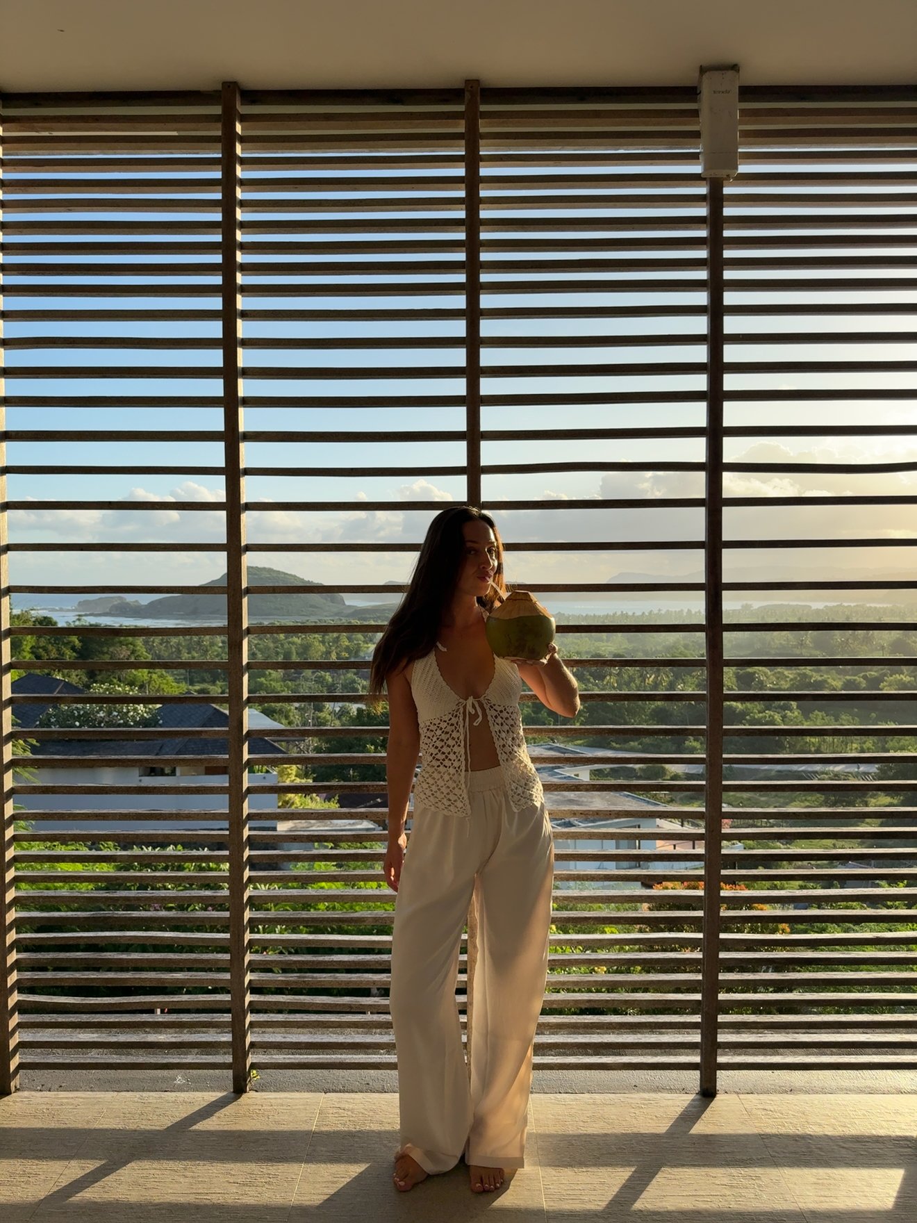 A woman stands in front of horizontal wooden slats, holding a coconut drink, with a scenic view of greenery and mountains in the background.