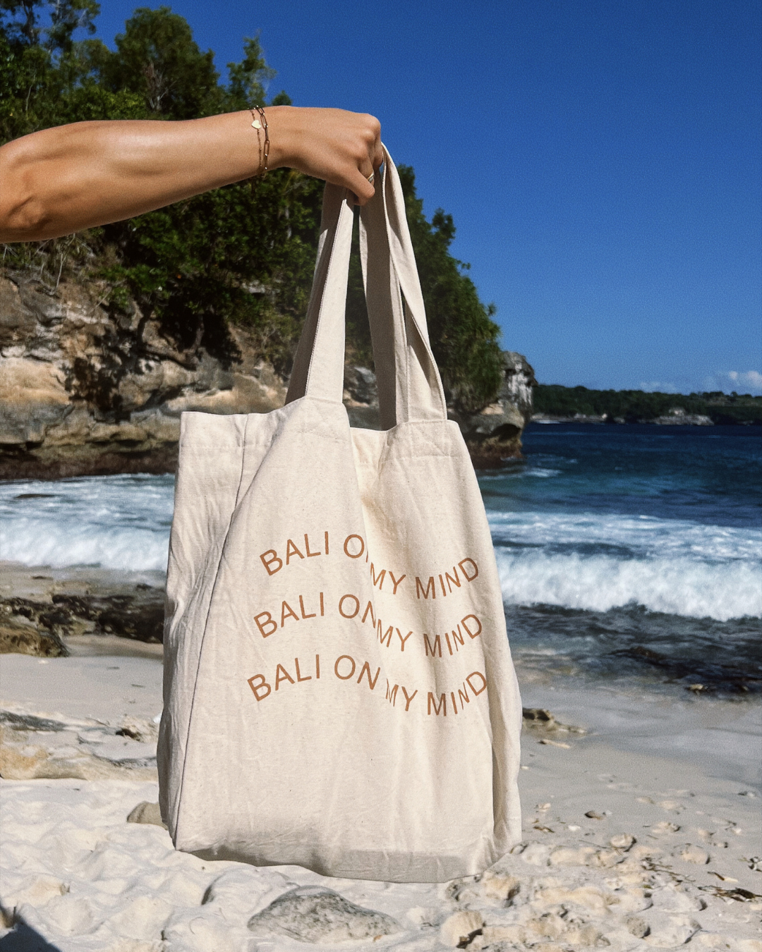 A person holding a beige tote bag with the phrase 'BALI ON MY MIND' printed on it, standing on a sandy beach with waves in the background.