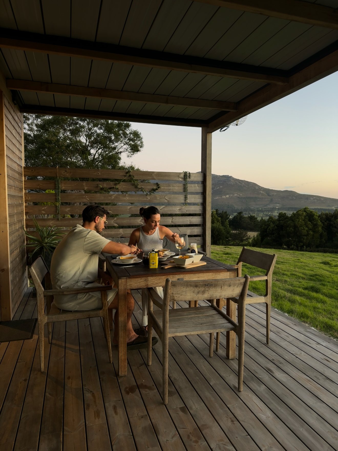 A couple enjoys a meal at a wooden table on a porch, with a scenic view of rolling hills and a sunset in the background.