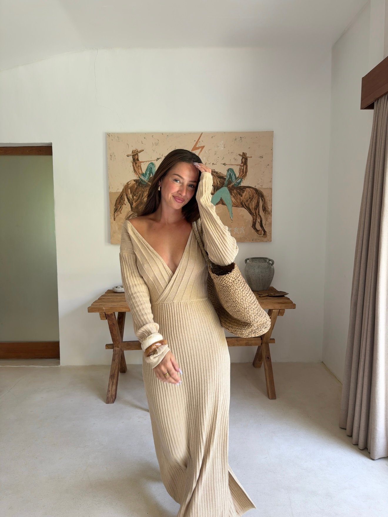 A woman in a beige ribbed dress poses confidently in a stylish indoor setting, with a wooden table and artwork in the background.