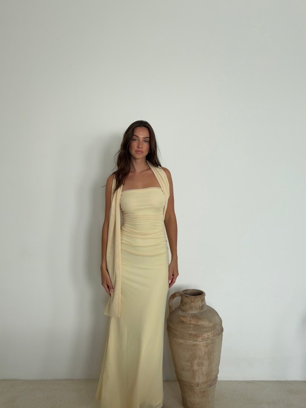 A woman stands elegantly in a fitted yellow dress next to a large pottery vase against a plain white wall.