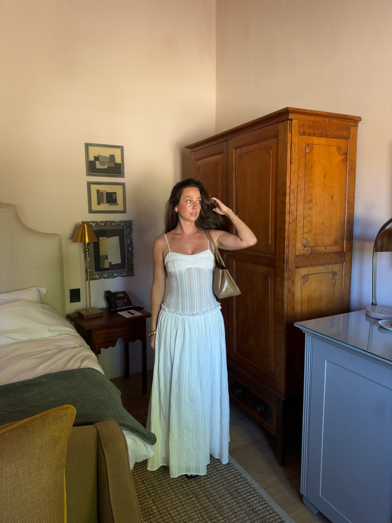 A woman stands in a stylish room, wearing a white dress and holding a small handbag. The background features a wooden wardrobe and a cozy bed.