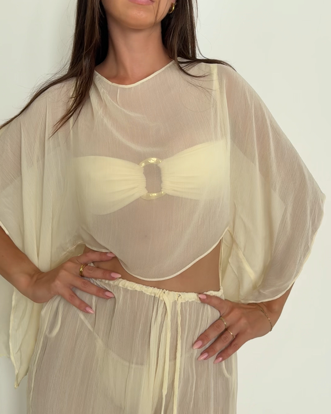 A model poses in a sheer, cream-colored outfit with a decorative buckle at the center, showcasing a stylish and elegant look.