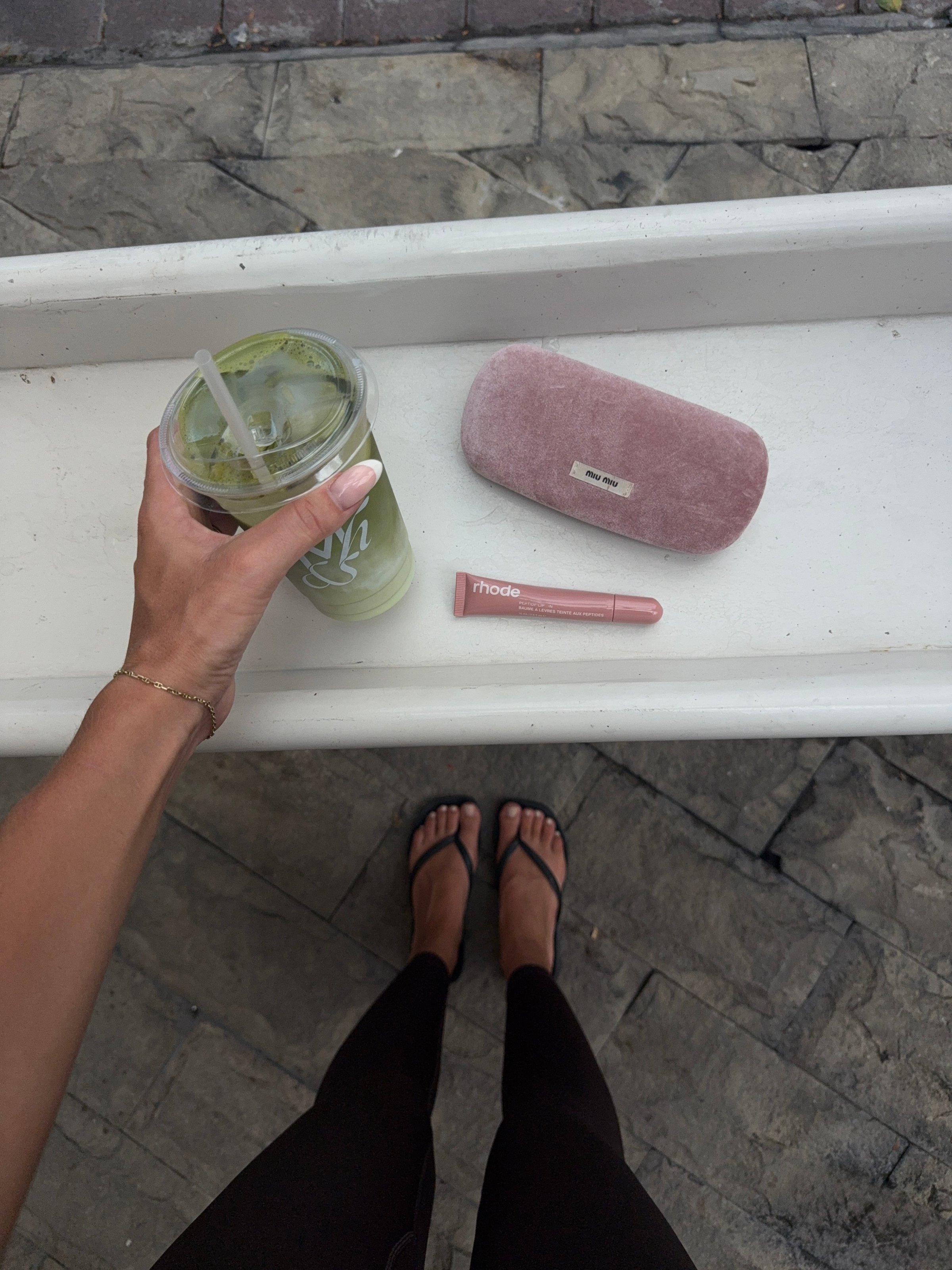 A person's hand holding a green drink, with a pink case and a beauty product placed on a white surface, while wearing black leggings and sandals.