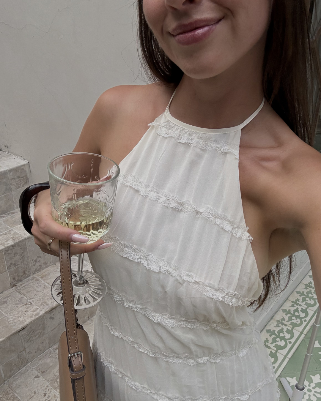 A woman in a white dress holds a glass of wine while posing for the photo, with a staircase and decorative tiles in the background.