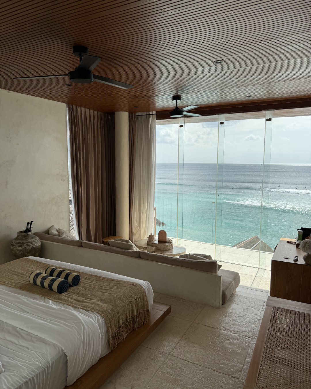 A luxurious bedroom with a view of the ocean, featuring a wooden bed, soft linens, and large glass windows that open to a serene seaside landscape.