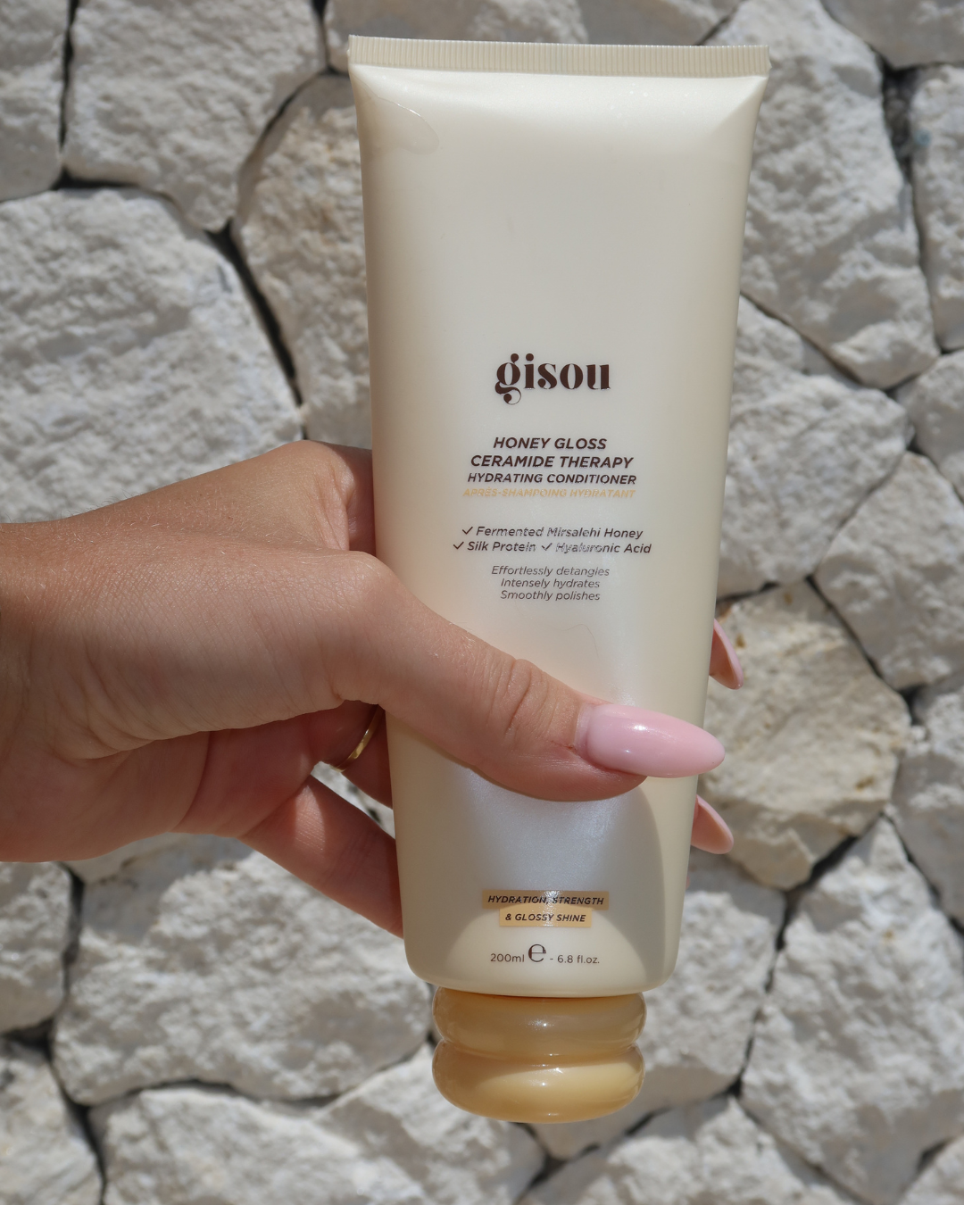 A hand holding a tube of Gisou Honey Gloss Ceramide Therapy Hydrating Conditioner against a textured stone wall.