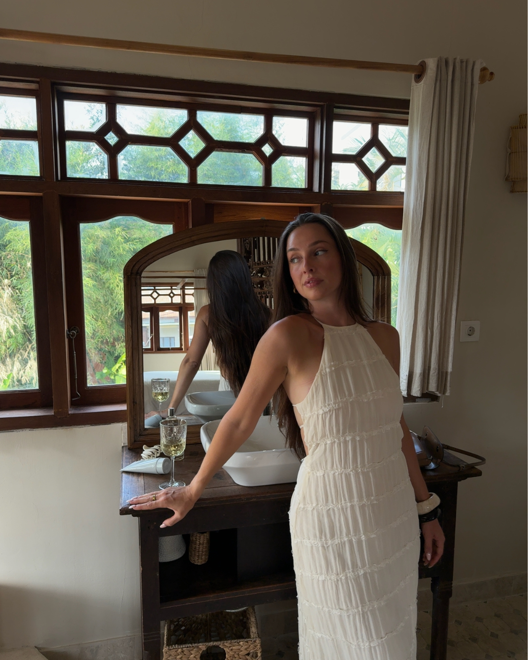 A woman in a white dress stands beside a wooden vanity with a mirror, showcasing a serene indoor setting with natural light filtering through the windows.