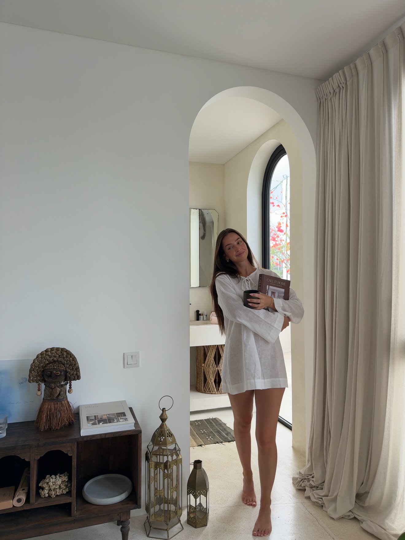 A woman in a white oversized shirt stands in a softly lit room, holding a book. The space features elegant decor with a lantern and a large window.