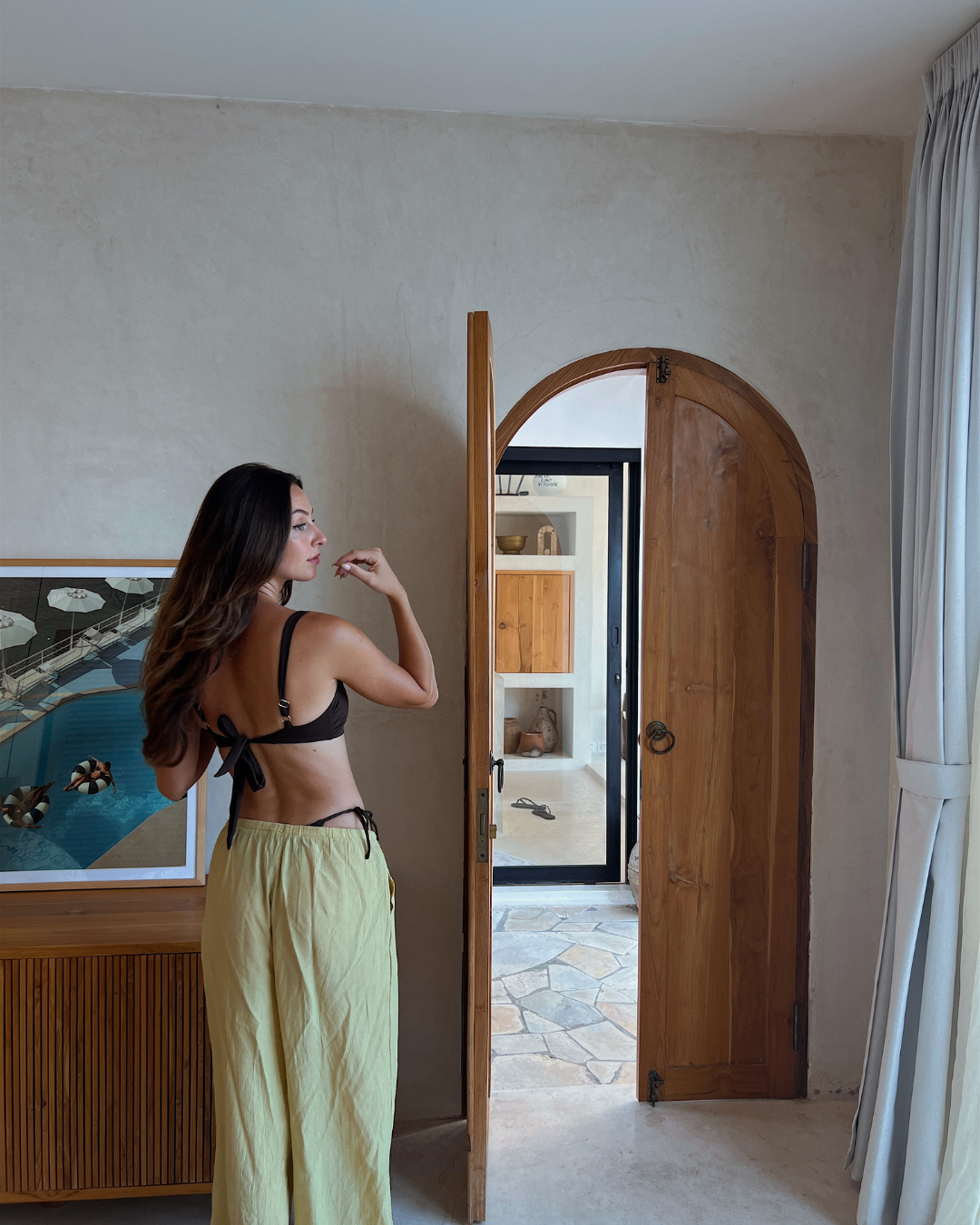 A woman in a brown bikini top and yellow pants stands with her back to the camera, looking towards an open door in a stylish interior space.