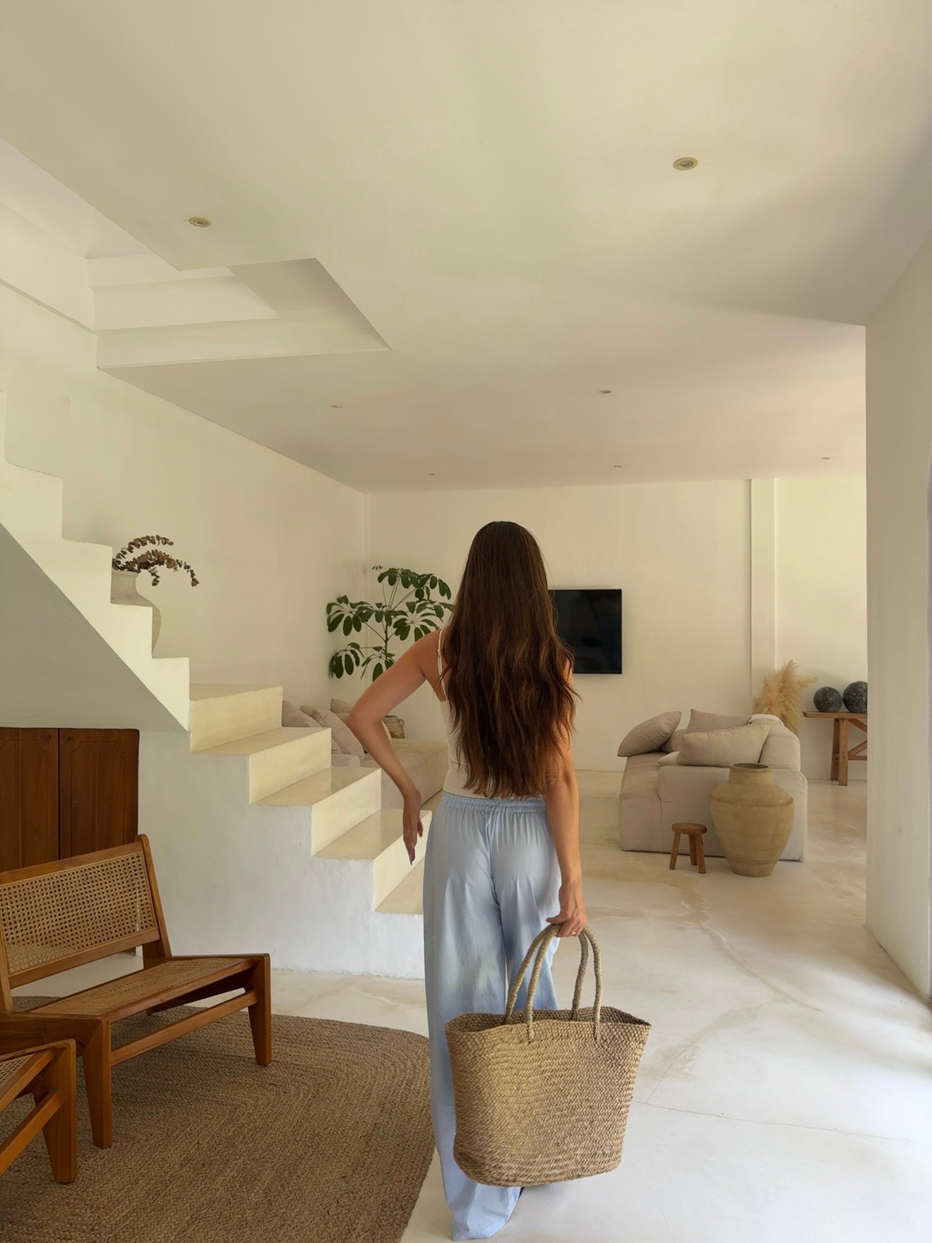 A woman with long hair walks away, carrying a woven bag, in a minimalist interior space with stairs and plants.
