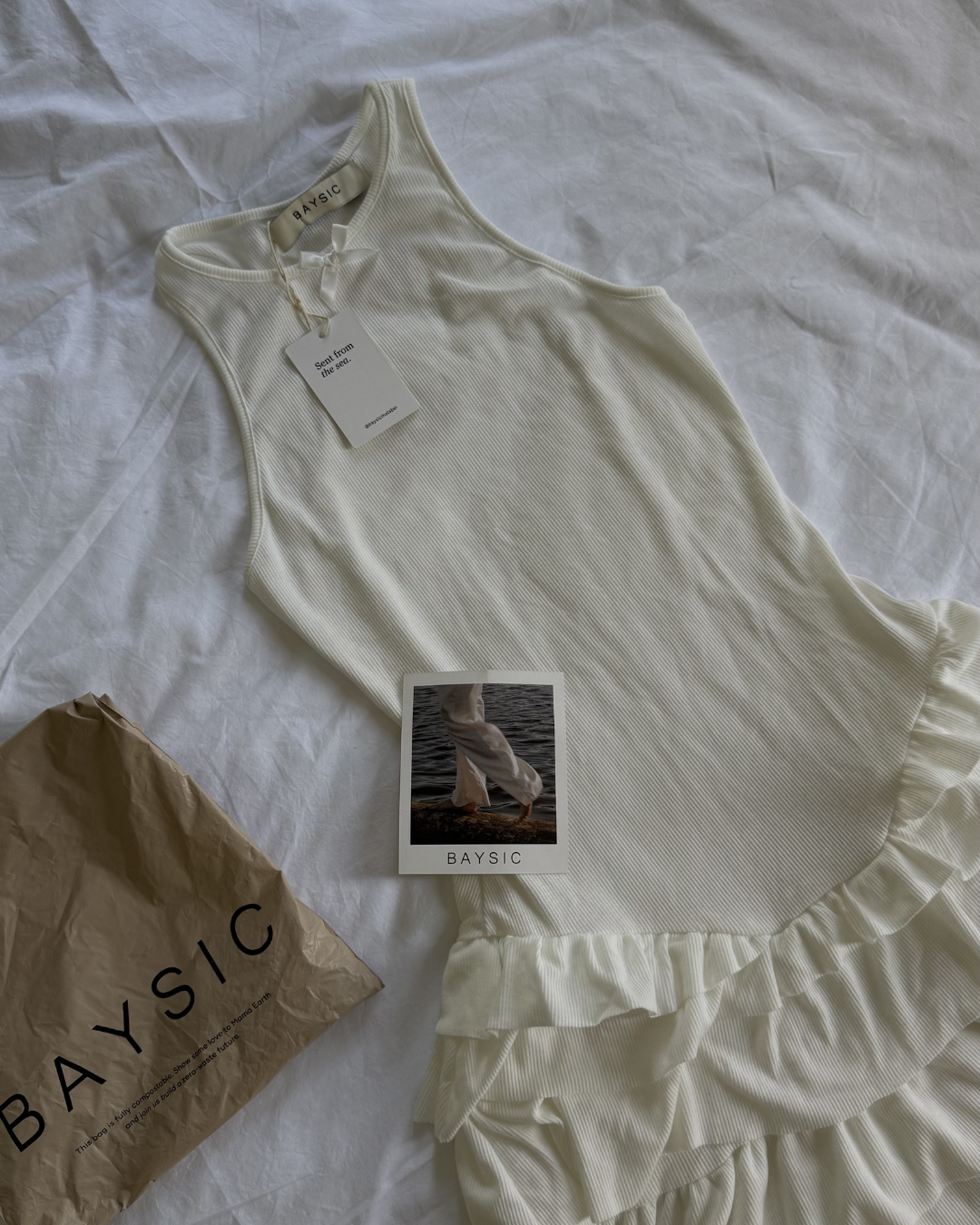 A light-colored dress with a ruffled hem laid out on a white surface, accompanied by a tag and a branded bag.