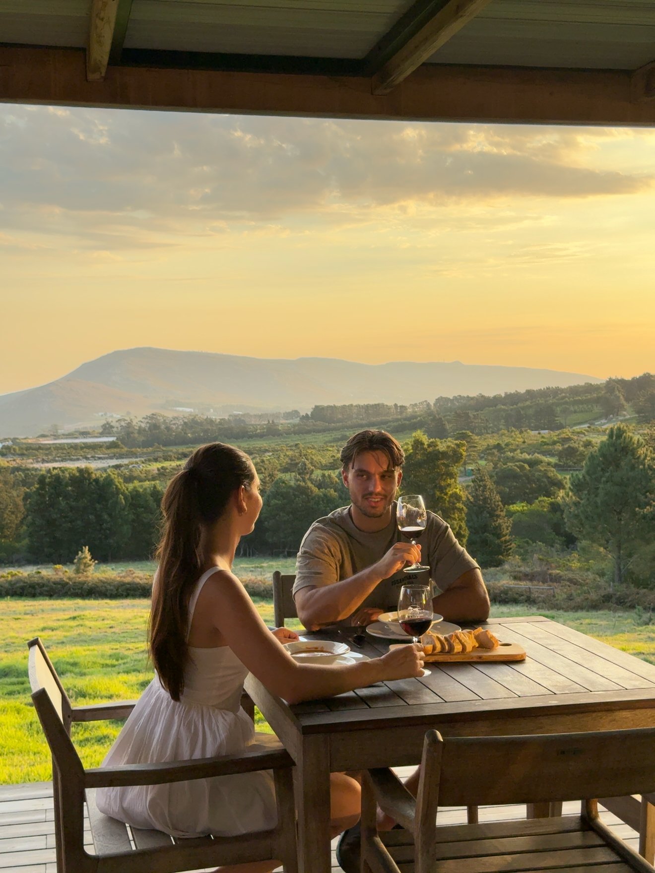 A couple enjoying a romantic dinner on a wooden patio, with a scenic view of rolling hills and a sunset in the background.