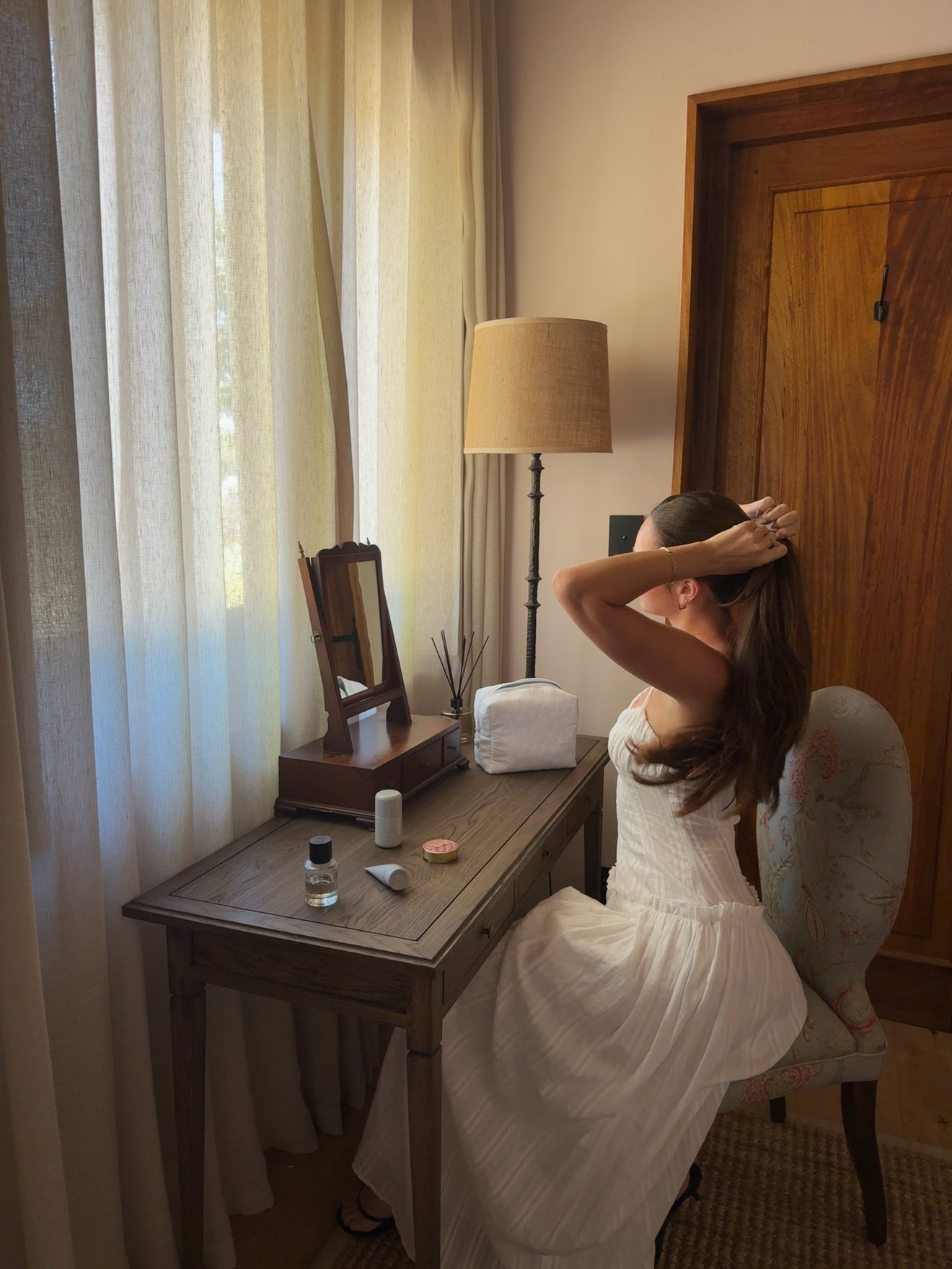 A woman in a white dress is sitting at a wooden vanity, tying her hair back in a well-lit room with sheer curtains.