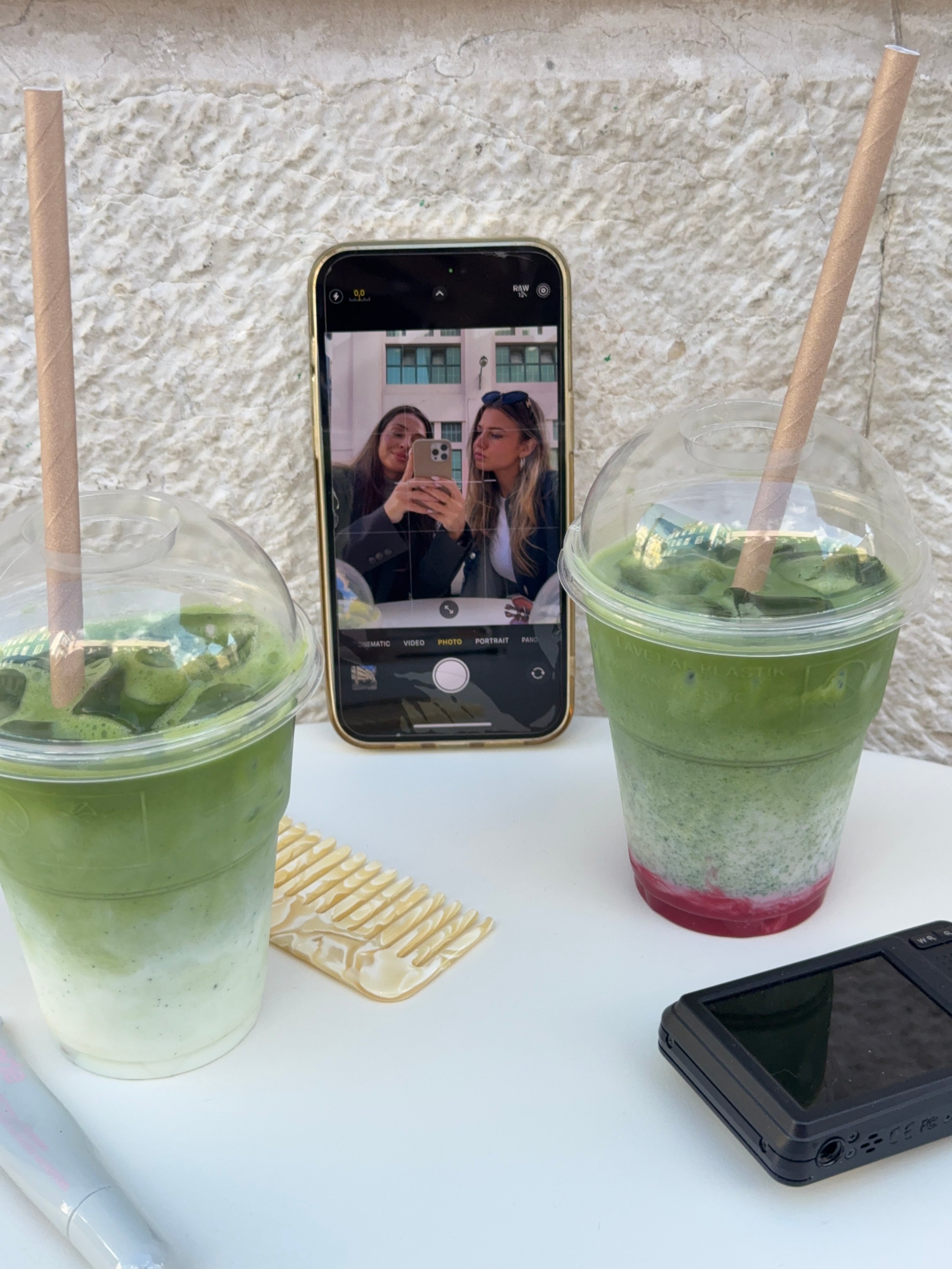 Two green beverages with layered colors are placed on a table, with a phone showing a selfie of two people in the background.