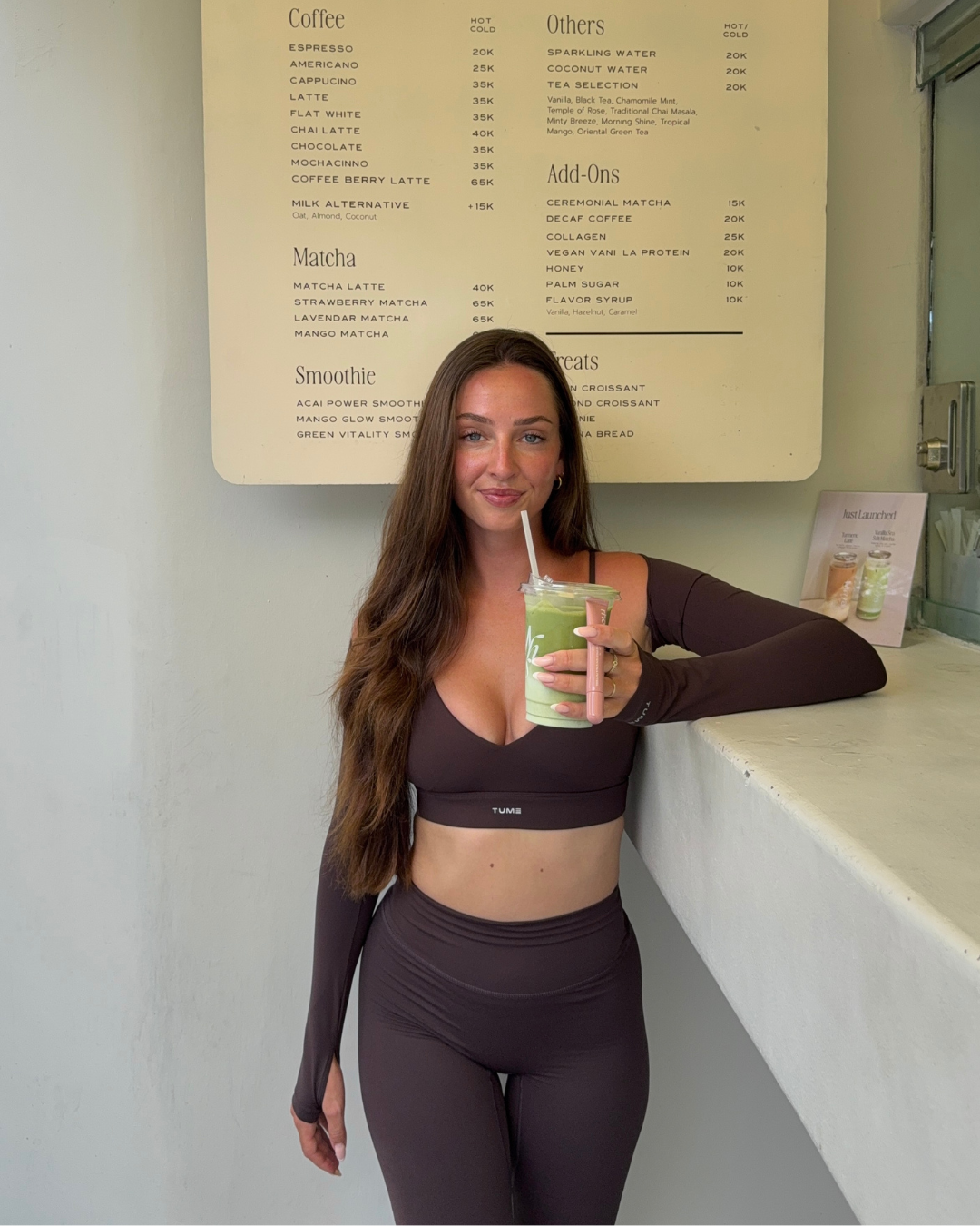 A woman in a brown crop top and matching leggings holds a green smoothie while standing at a café counter with a menu in the background.