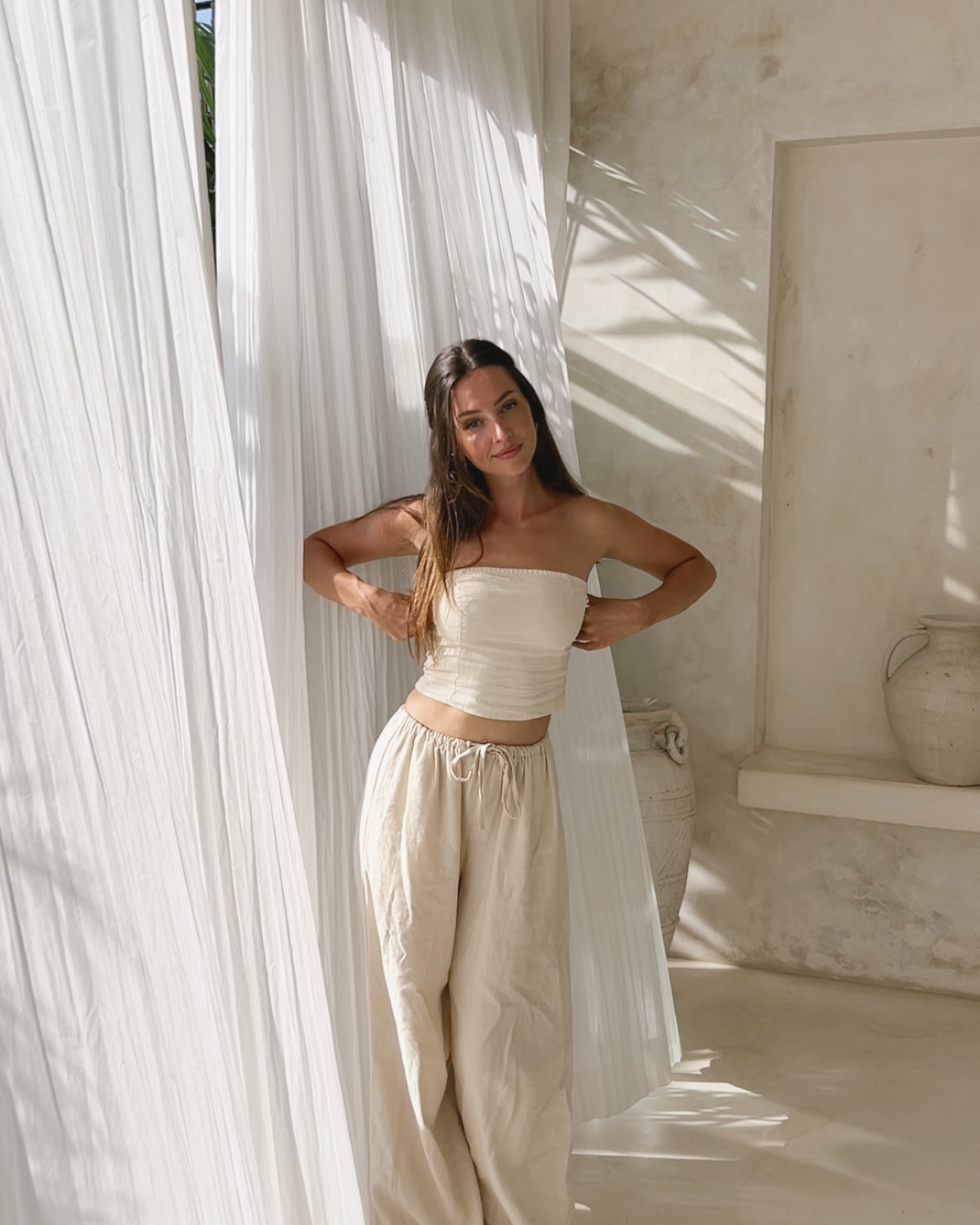 A woman stands gracefully in a sunlit room, wearing a strapless top and loose-fitting pants, with sheer white curtains in the background.